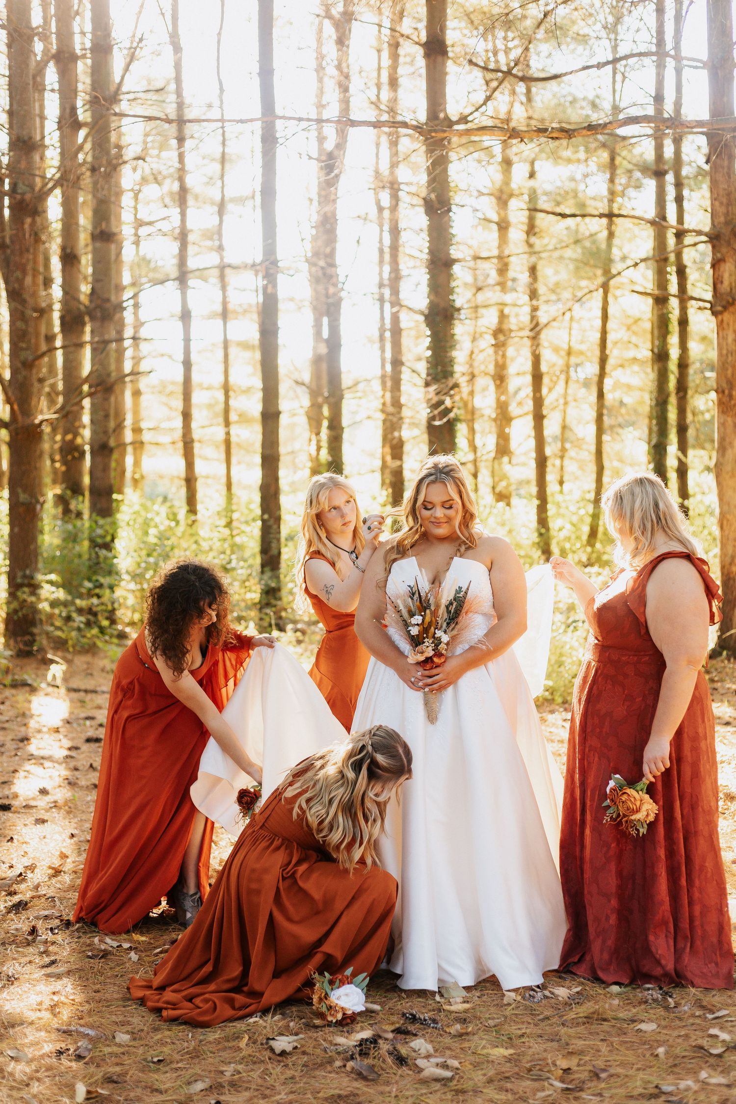 Bridesmaids in burgundy dresses pose with bride in a sunlit forest setting.