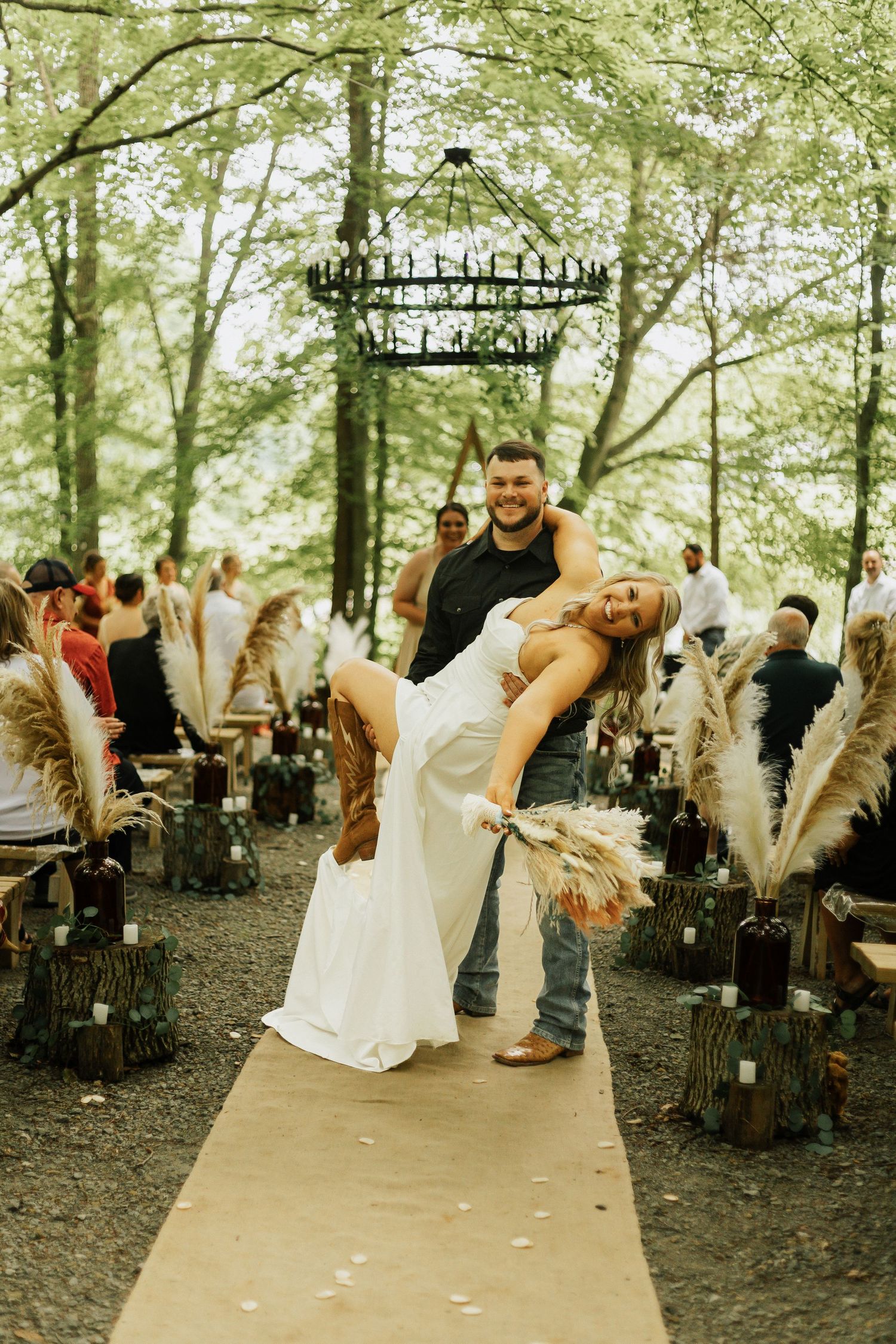A couple dances together on a rustic outdoor aisle lined with guests and hanging chandeliers.