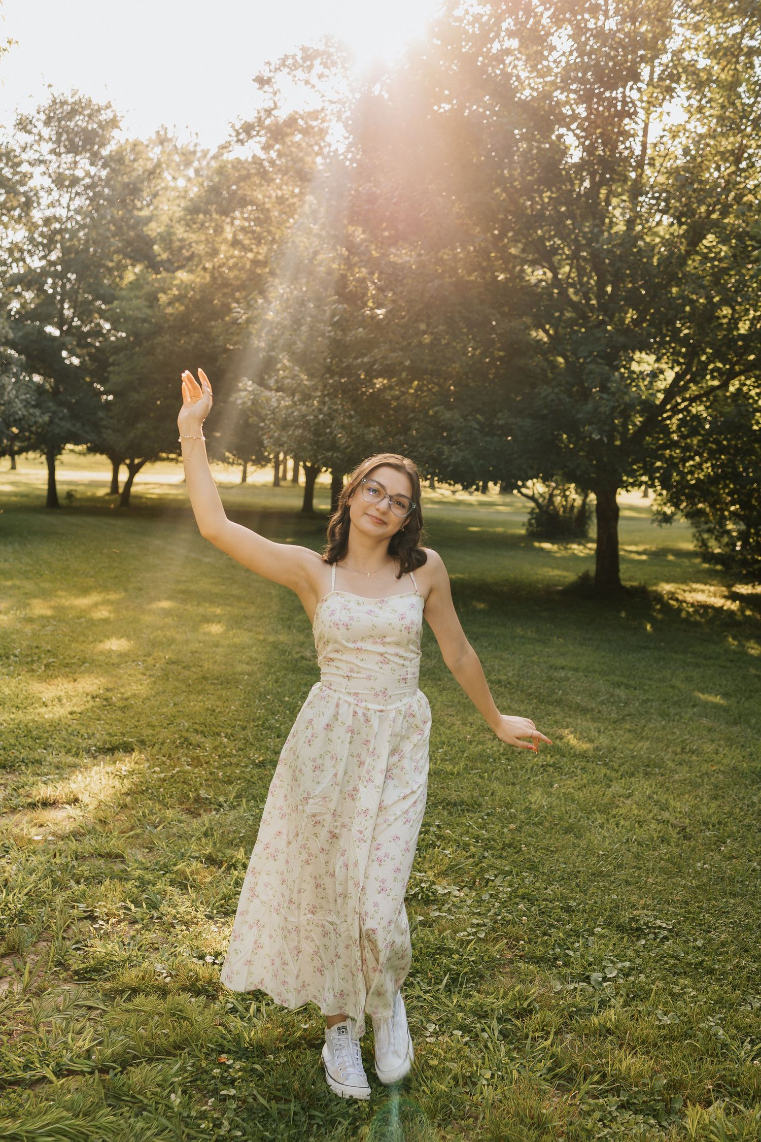 Young woman in floral midi dress and glasses dancing in sunlit park with lush green trees in background.