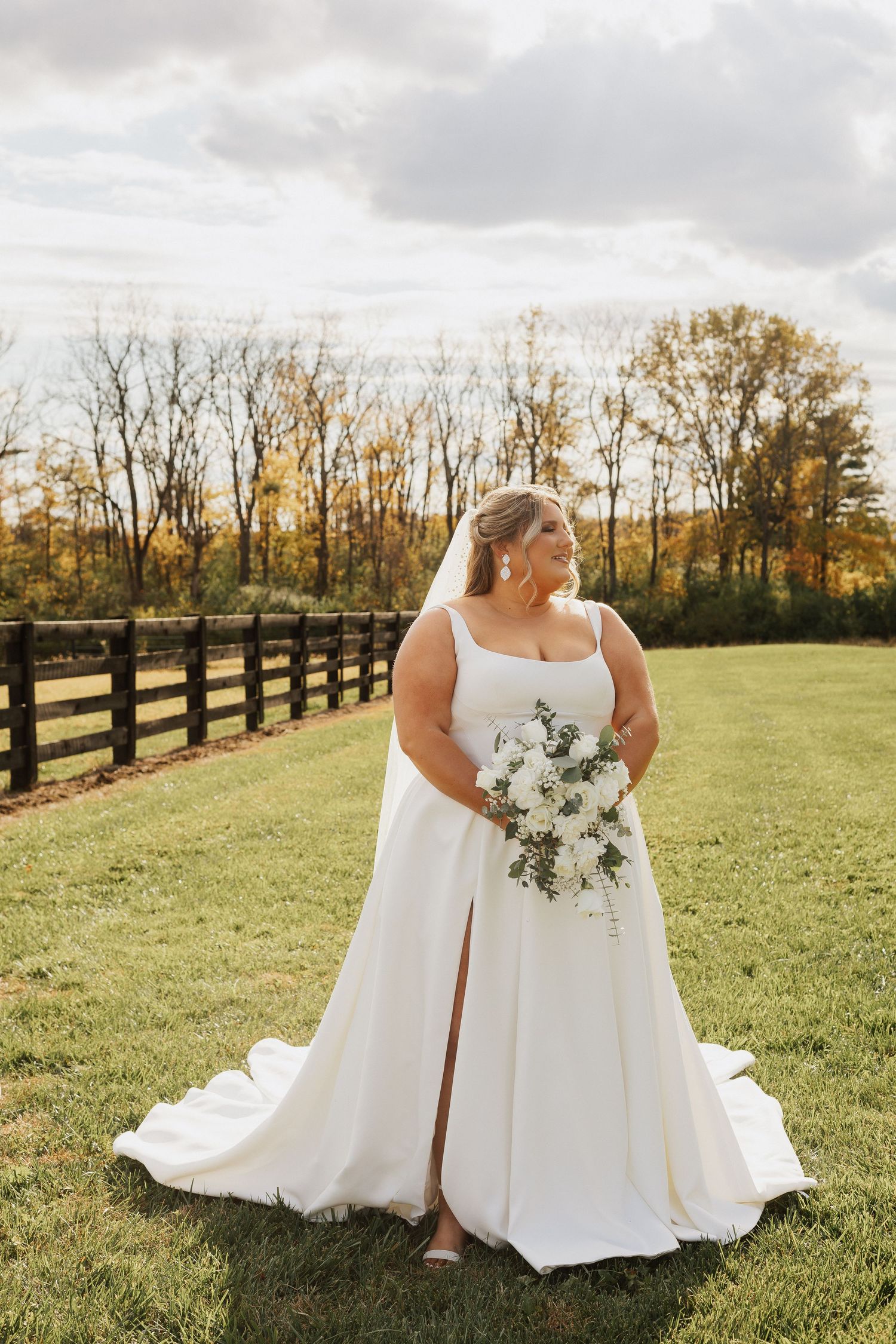 Bride in elegant white dress poses with bouquet in front of rustic wooden fence.
