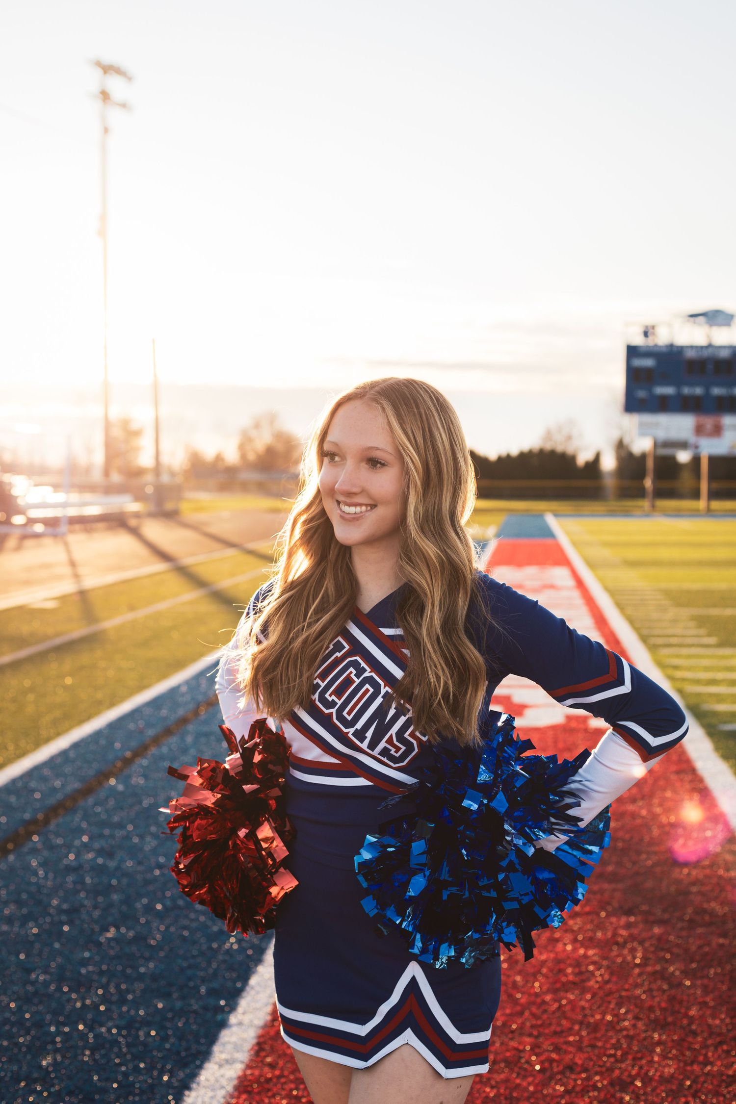 Cheerleader in navy uniform poses with red and blue pom poms on athletic field.