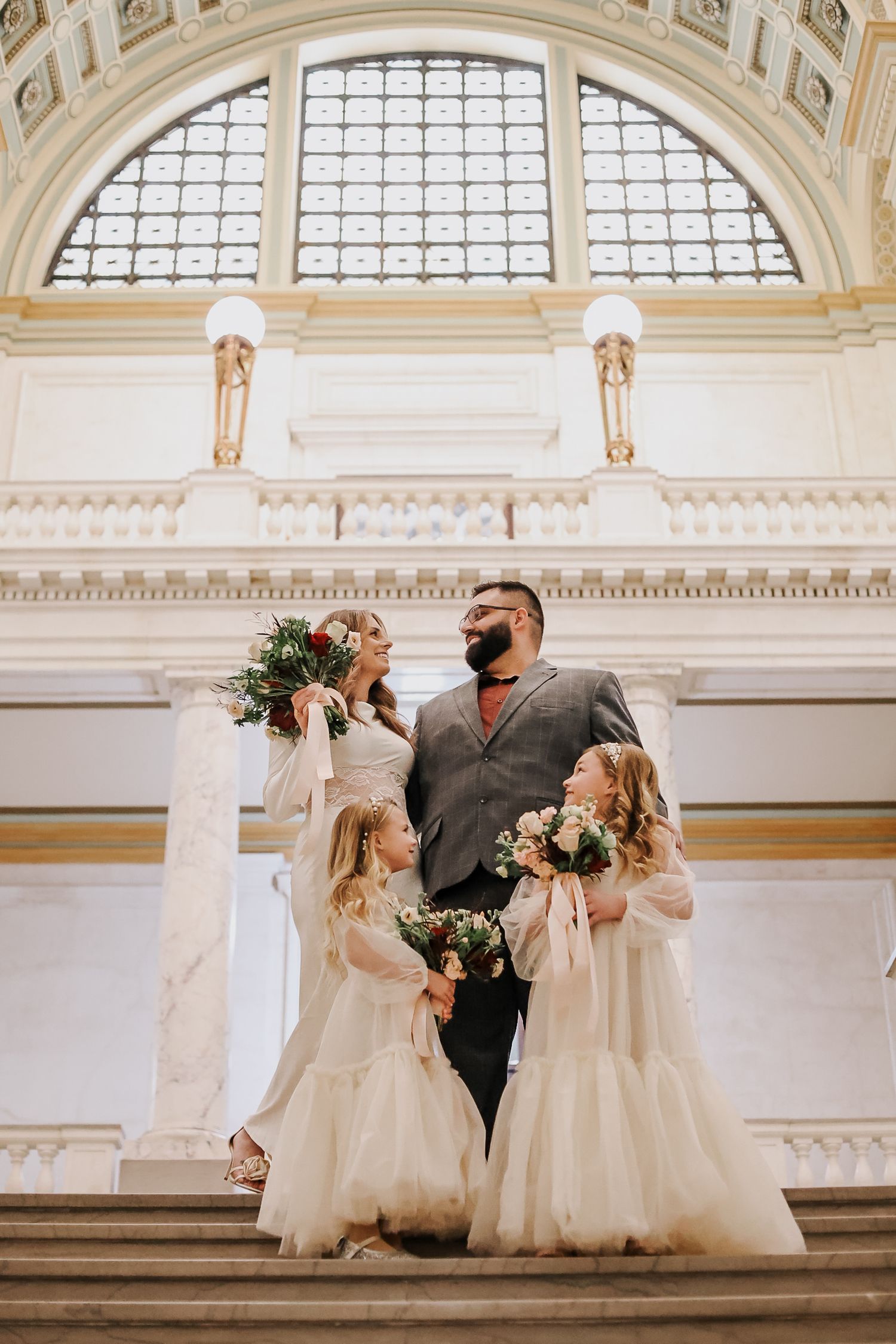 A wedding group seated beneath grand arched windows in an ornate architectural setting.