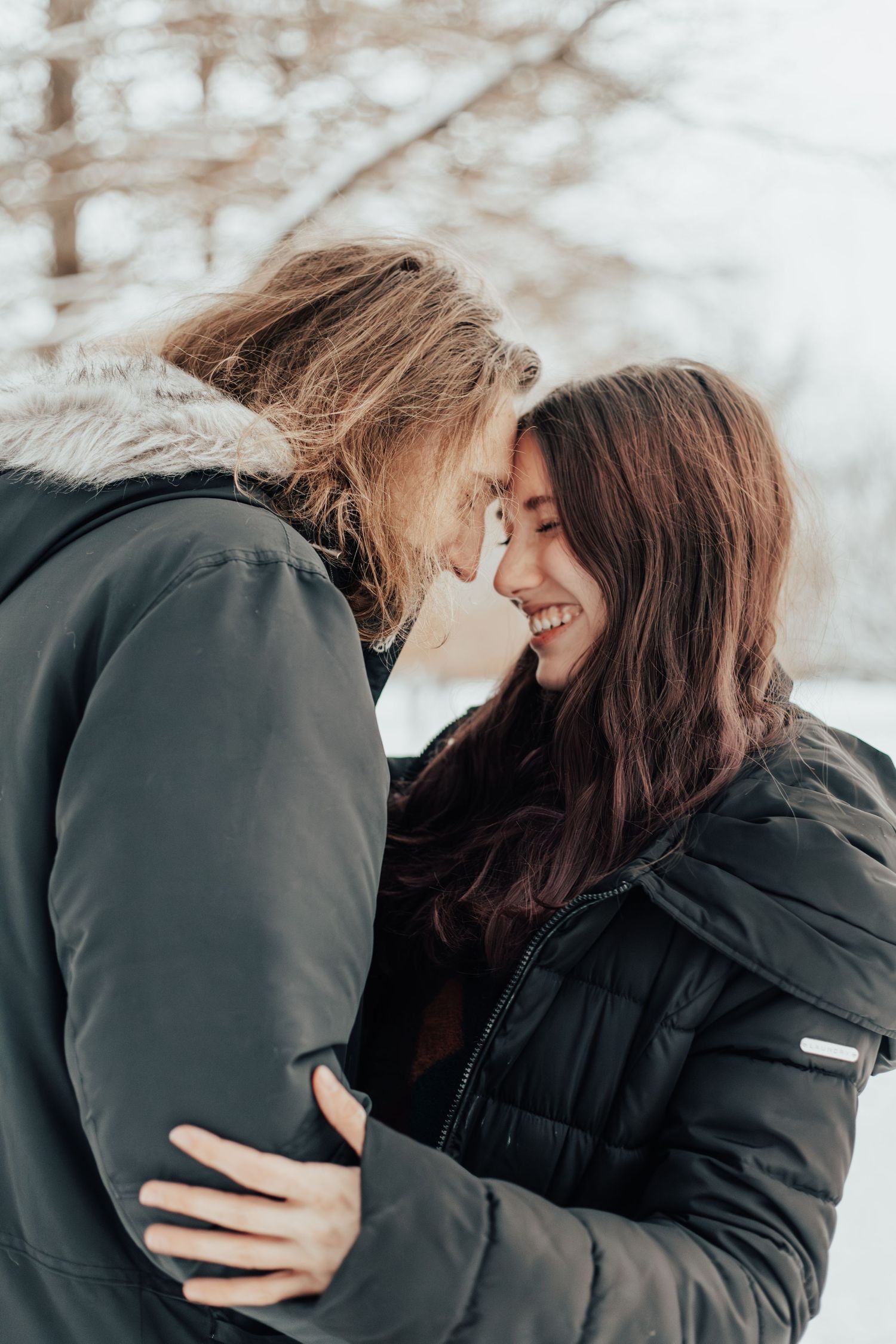 A tender moment between a couple in winter coats against a snowy backdrop.