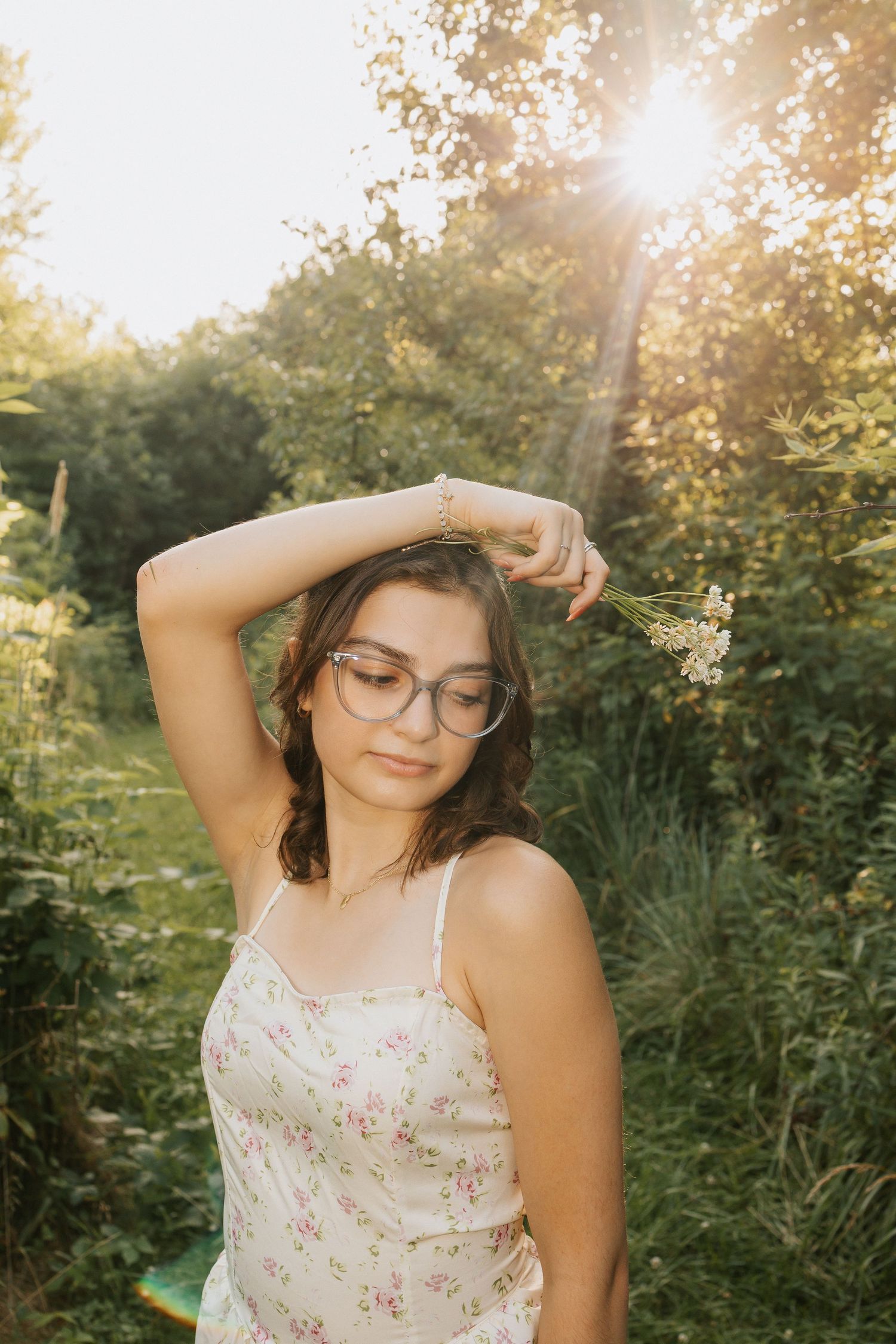 Young woman in floral sundress and glasses holds wildflowers in golden hour sunlight surrounded by lush green foliage.