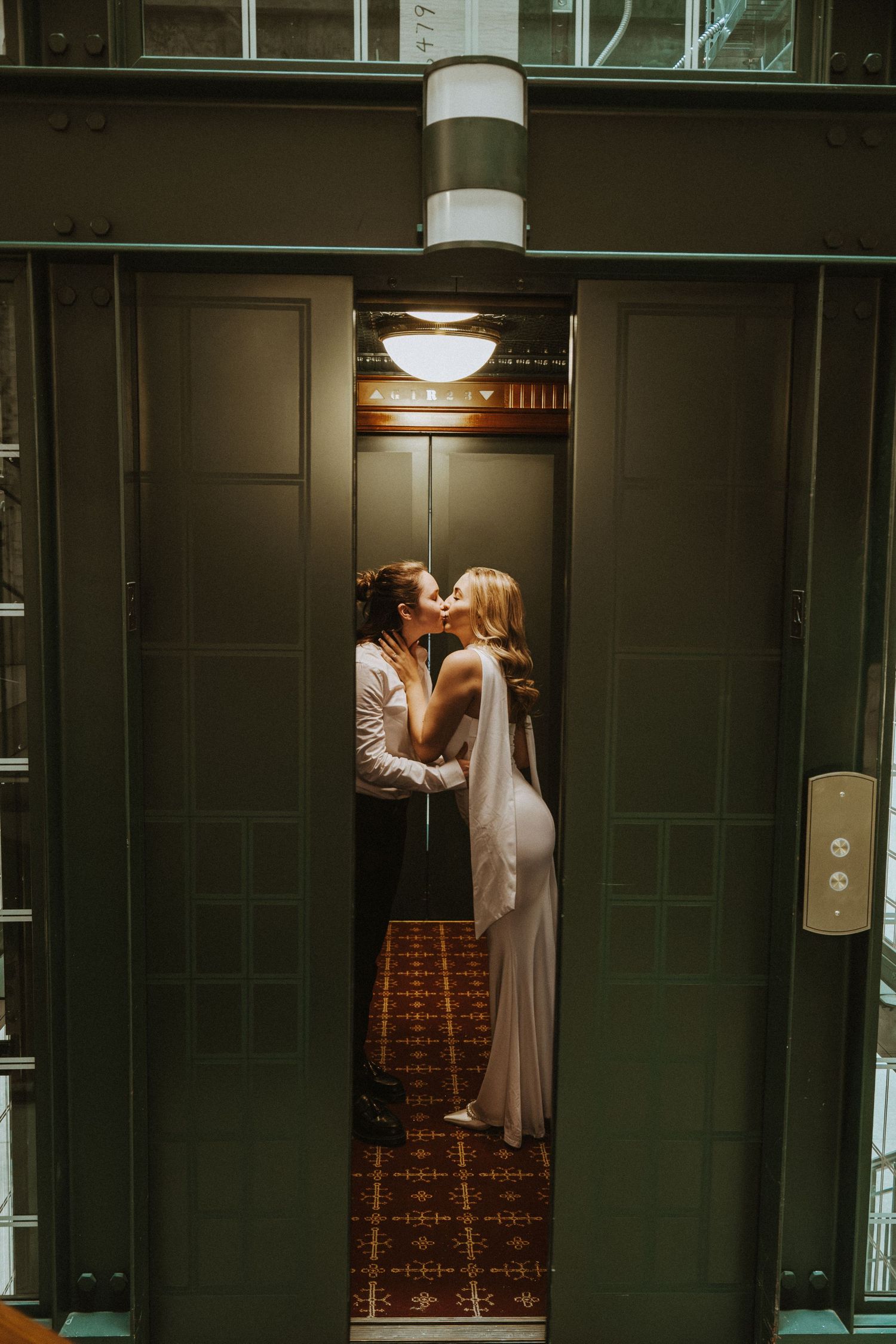 Couple sharing a romantic kiss in an elegant elevator doorway, bride in white gown with veil, groom in white shirt.