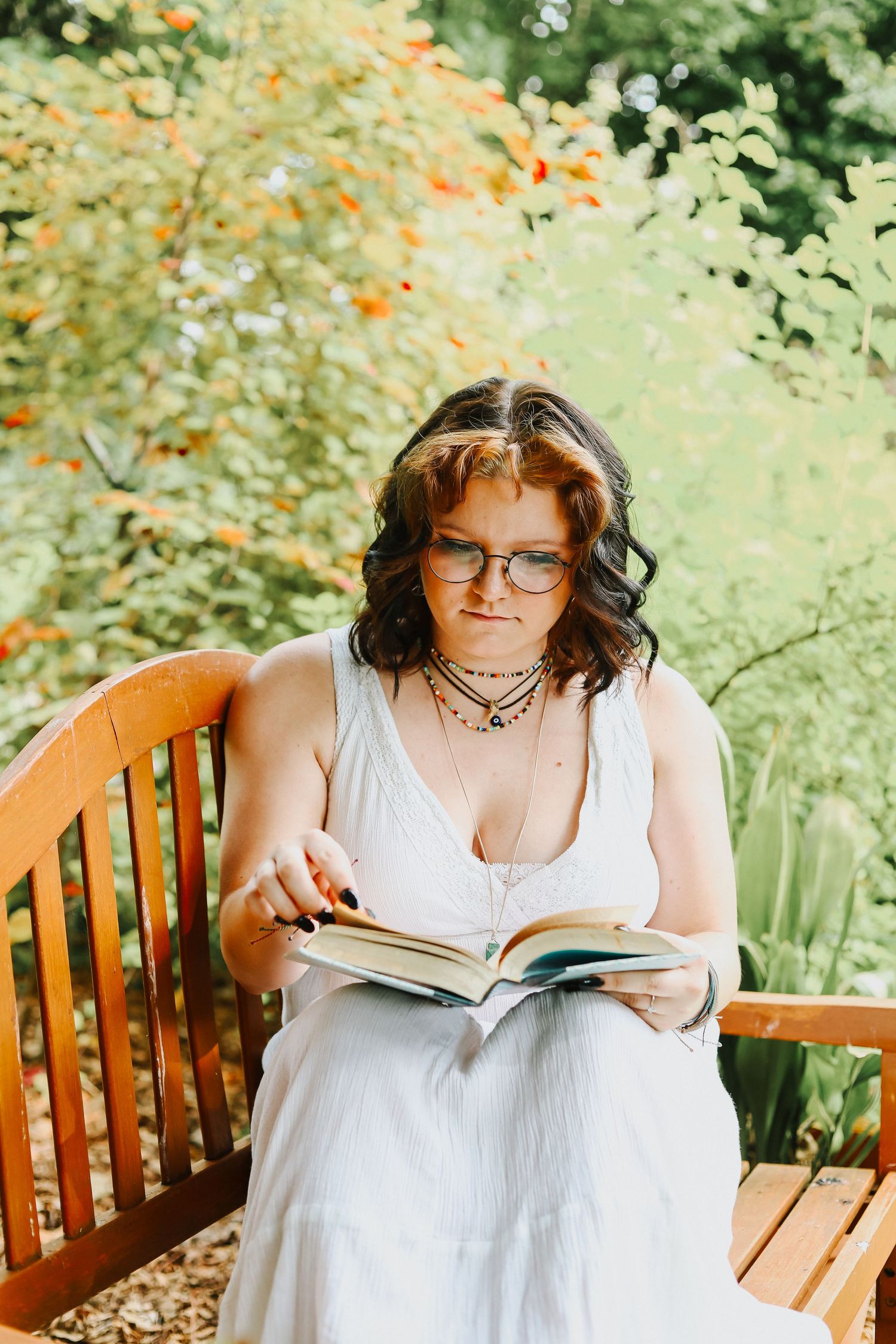 Person in white dress reading a book while relaxing on a wooden bench in a lush garden setting.