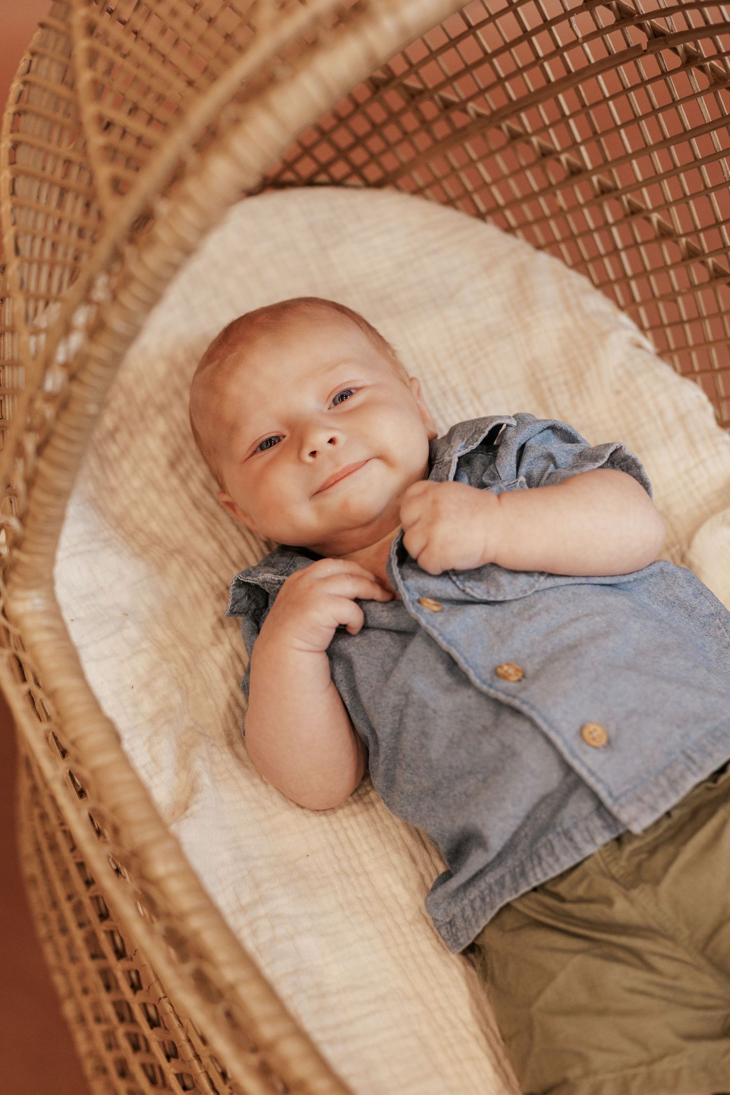 Baby in blue outfit lying in woven rattan chair with cushioned interior.