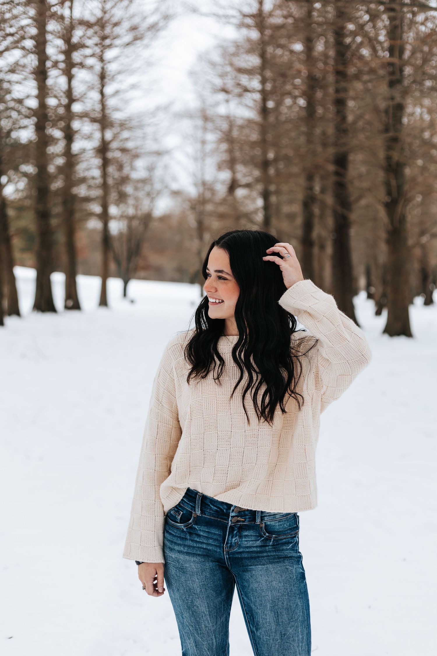 Person in cream sweater and jeans posing playfully in snowy winter forest setting.