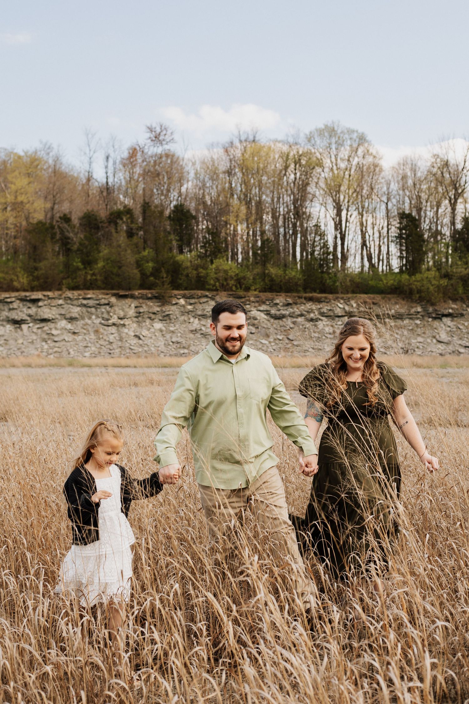 Family walking through tall golden grass with rocky cliff and trees behind them.