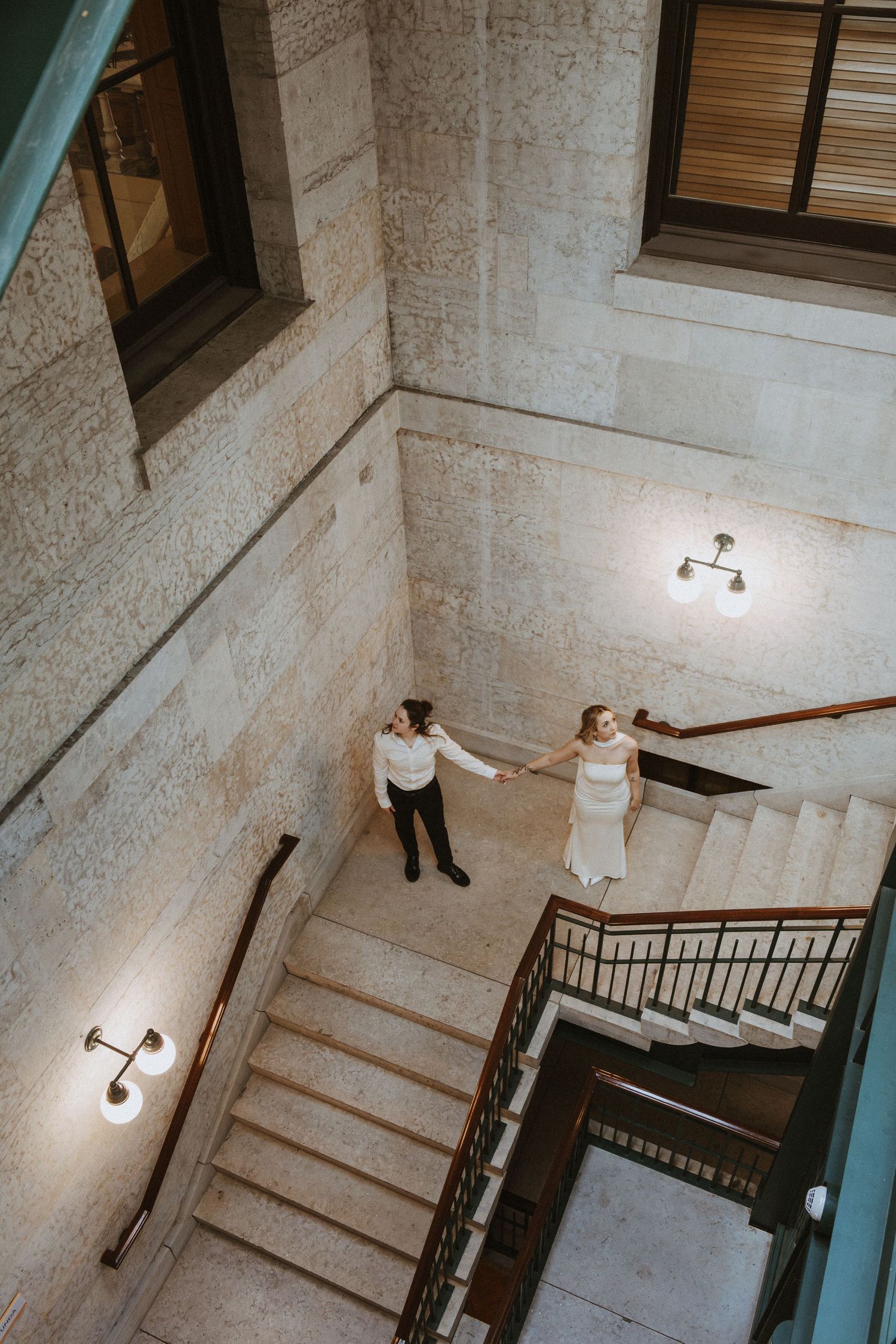 Bride and groom hold hands on elegant marble staircase, viewed from above in a grand interior venue.