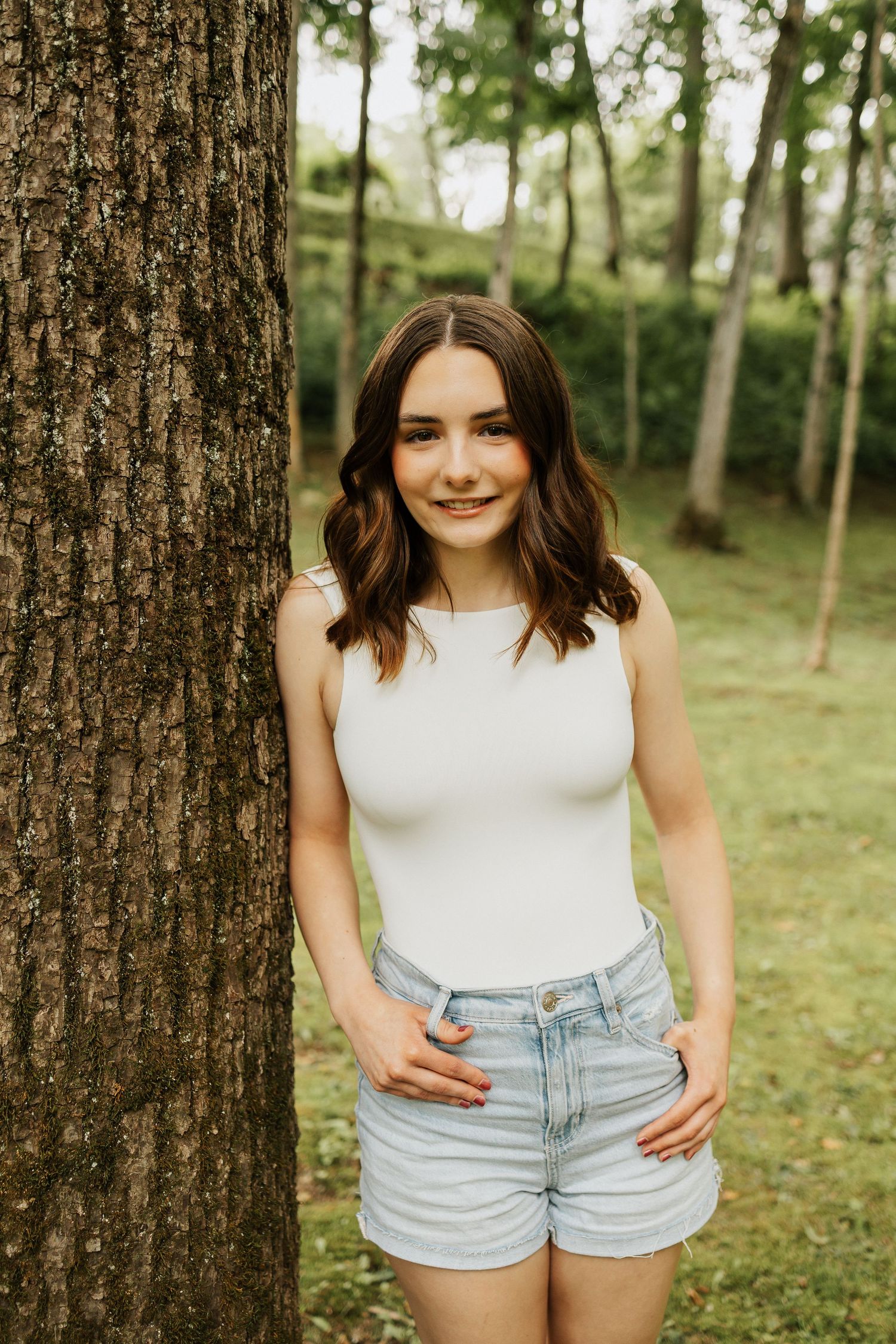 Young woman in white tank top and denim shorts smiling beside a tree in a lush green forest setting.