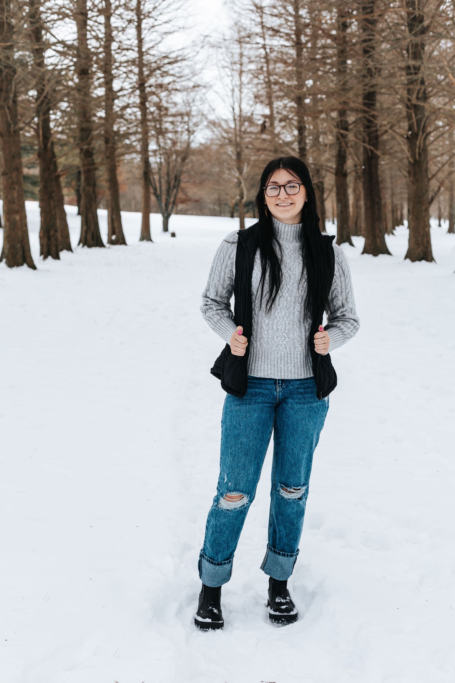 Person in grey sweater and ripped jeans standing in deep snow among bare trees.