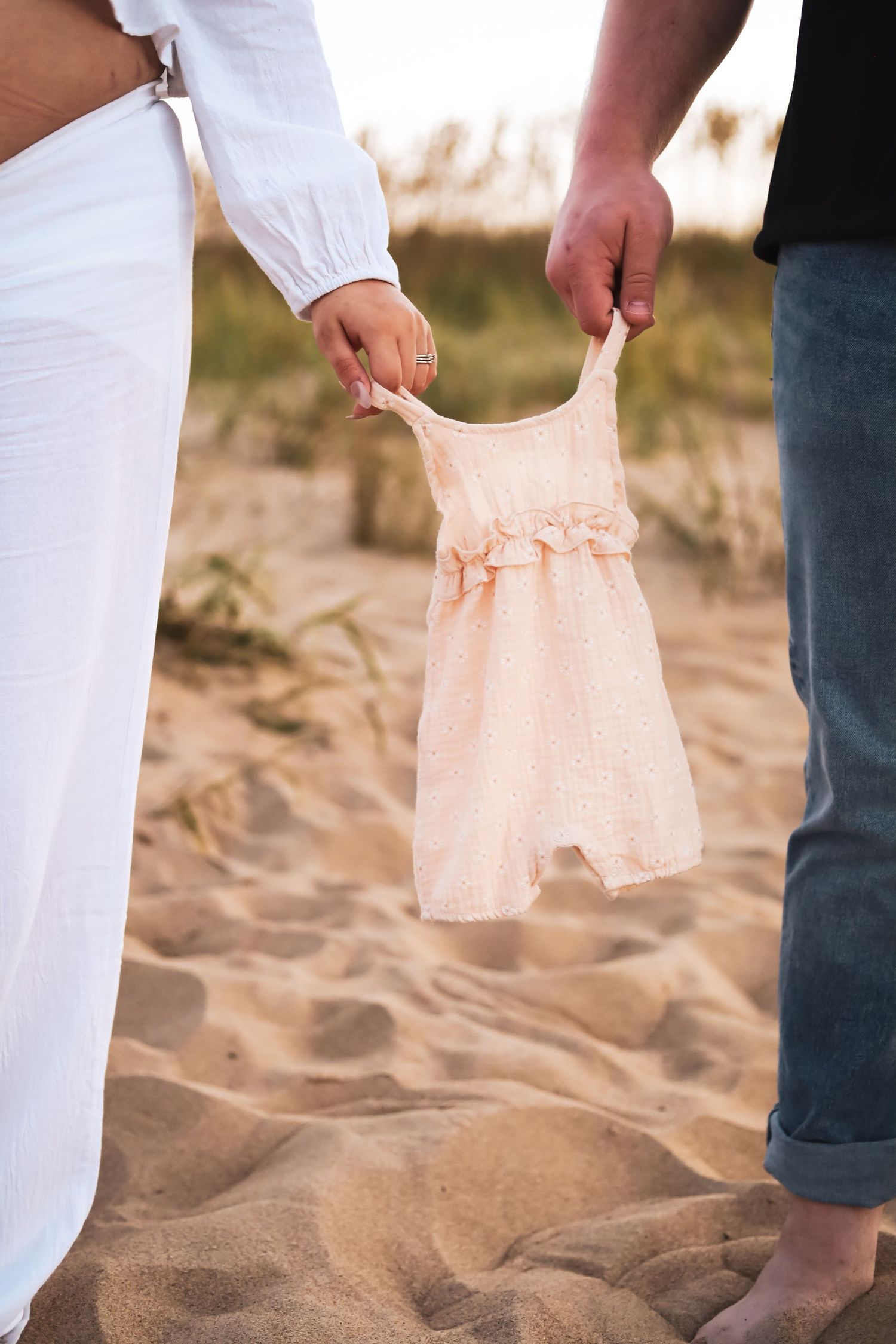 Hands holding tiny pink baby clothes on sandy beach during sunset.