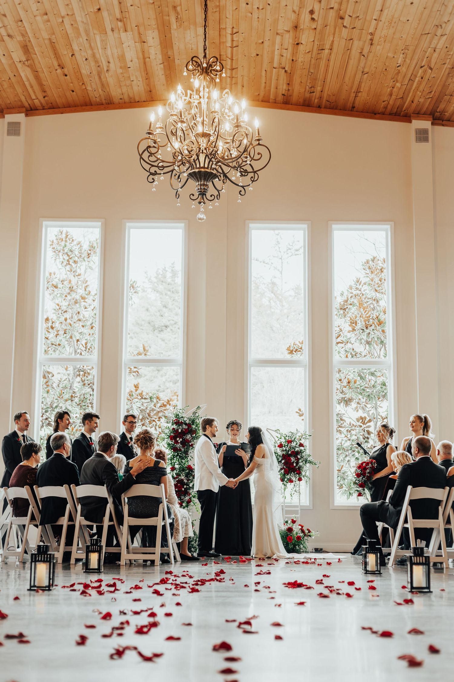 Wide shot of an elegant wedding ceremony space with wooden ceiling, chandeliers and scattered rose petals.
