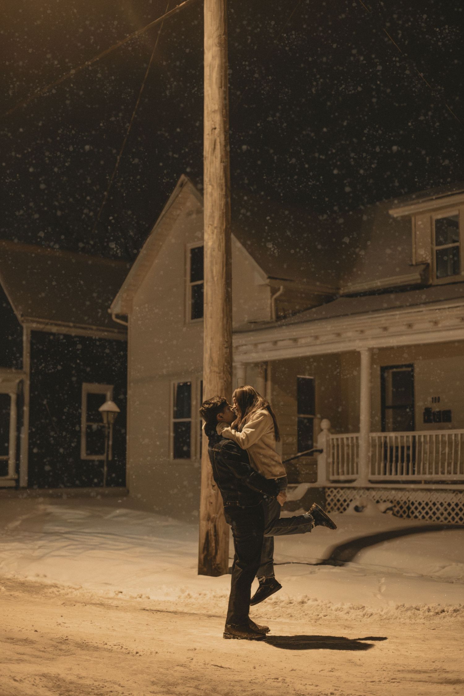 A snowy nighttime scene with white houses and falling snow illuminated by outdoor lighting.