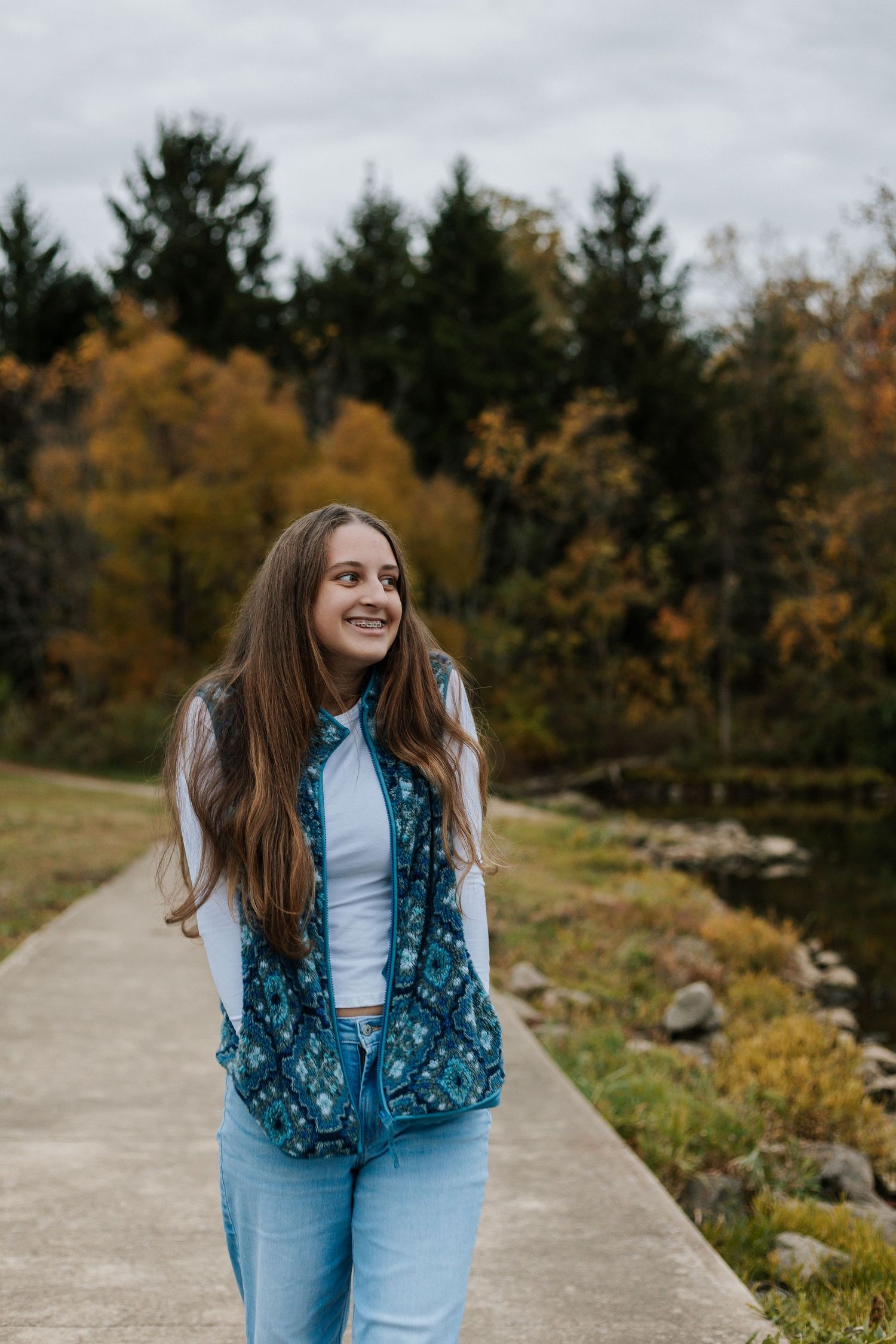 Person walking along lakeside path surrounded by fall foliage and evergreen trees.