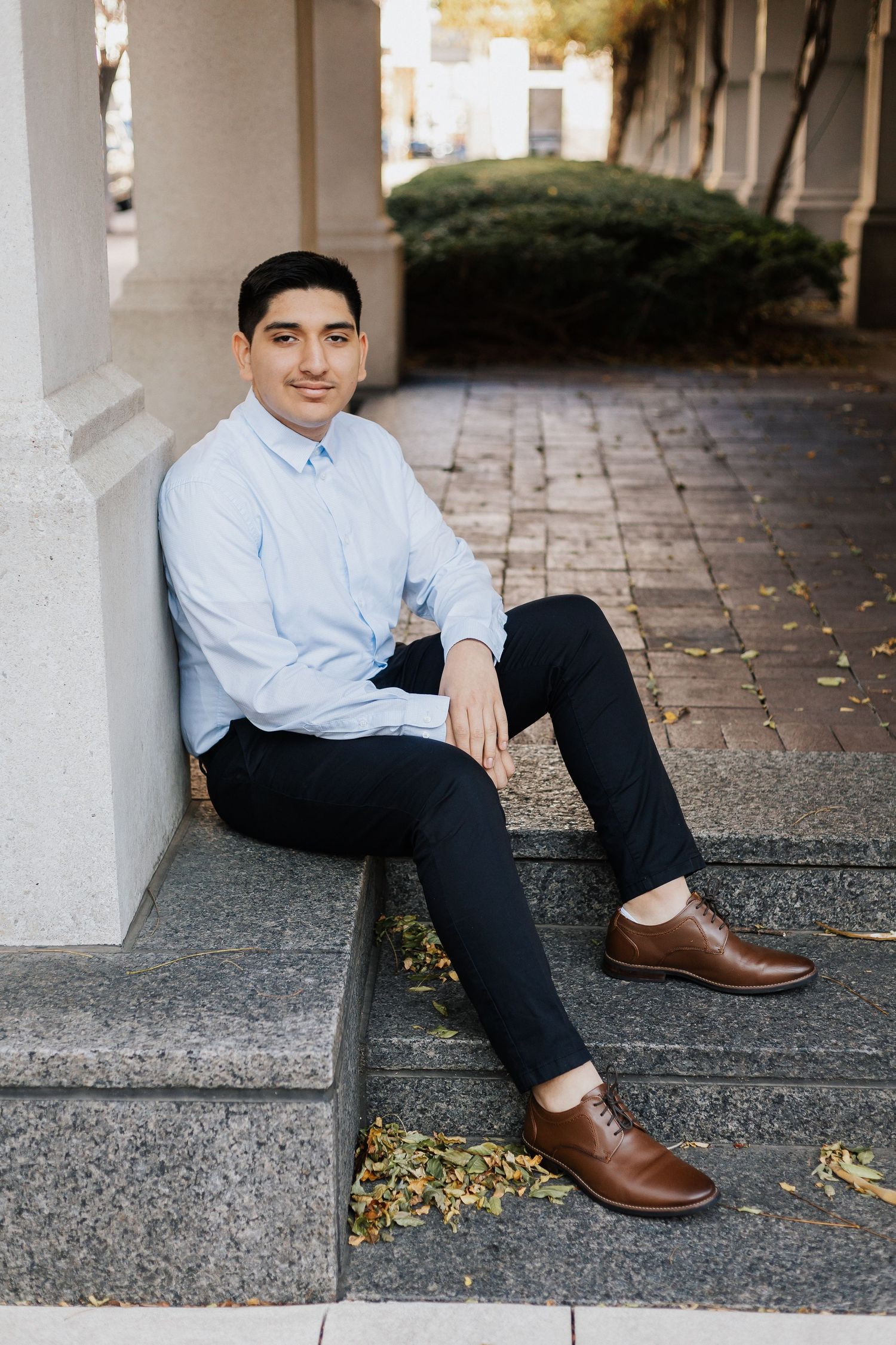Casual business portrait showing brown leather shoes and relaxed seated pose against stone columns.
