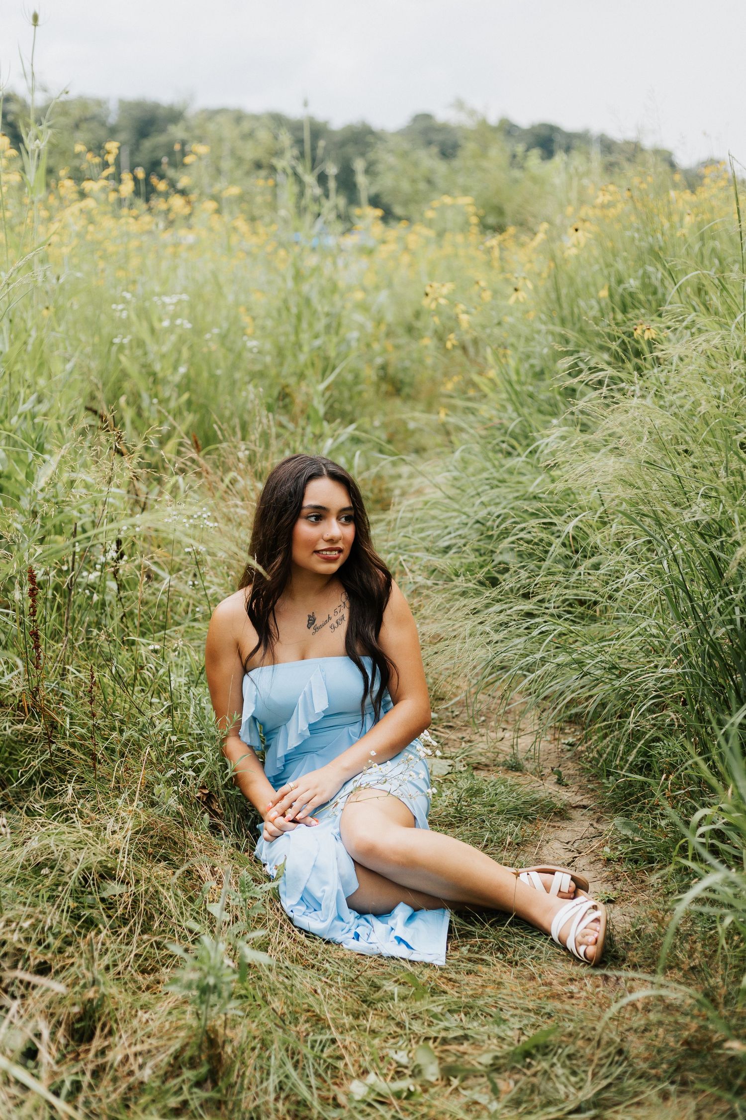 Person in light blue dress poses sitting among tall wildflowers and grass.