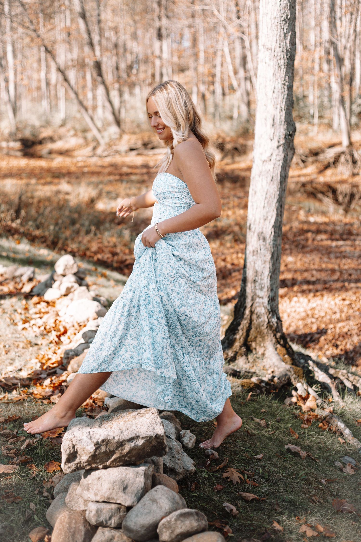 Someone in a blue dress poses gracefully on rocks by a wooded pond.