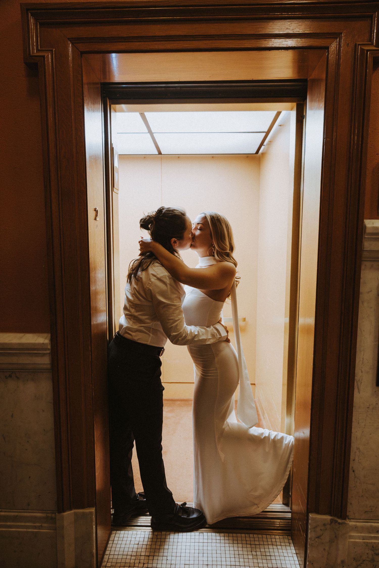 Two brides share a romantic kiss in a vintage elevator doorway, one in a white gown and the other in a white shirt and black pants.