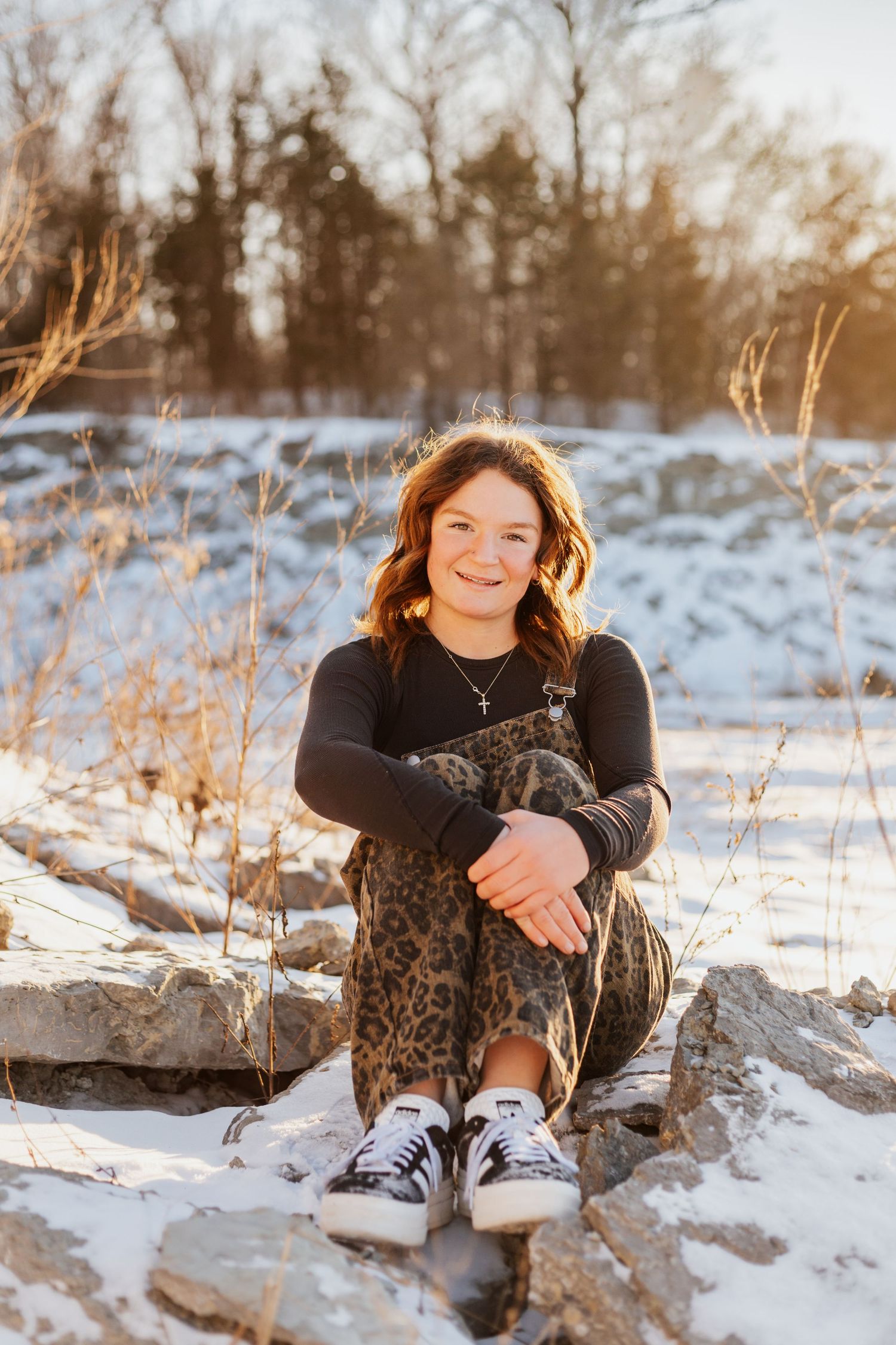 Person in black sweater and leopard dress sits on snowy rocks at sunset.