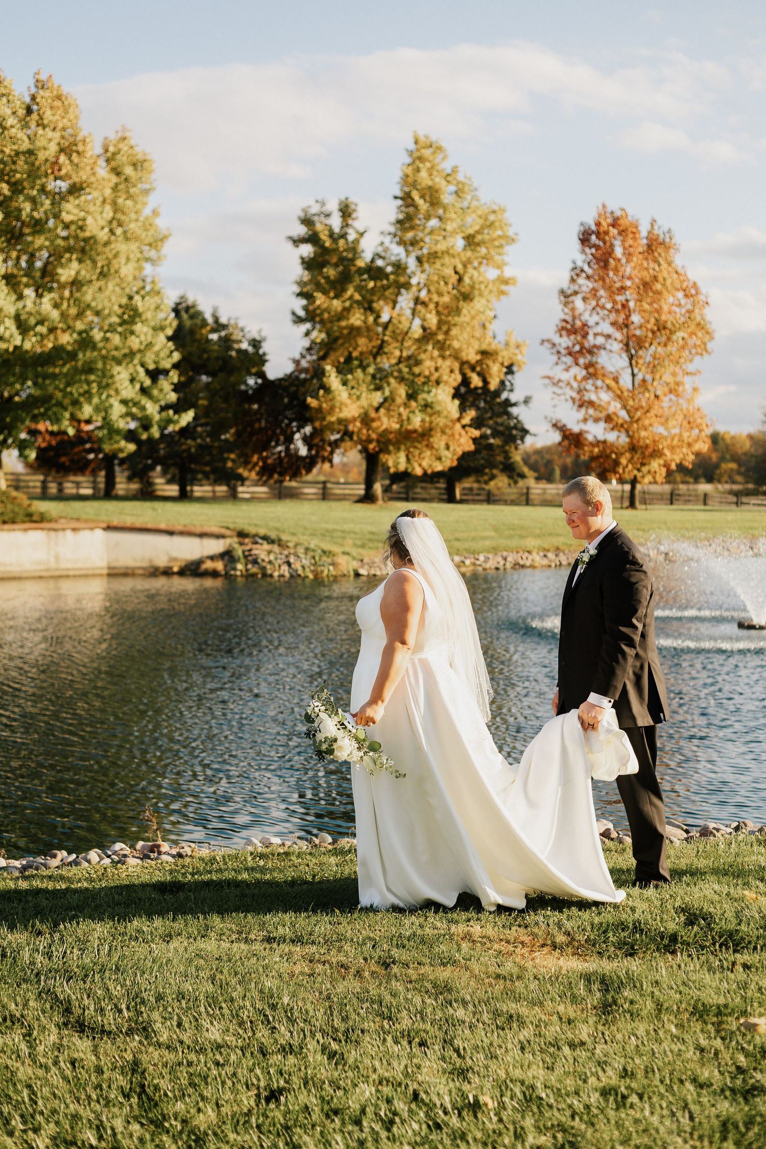 Newlyweds walk along a serene lakeside path during golden hour with autumn trees in background.