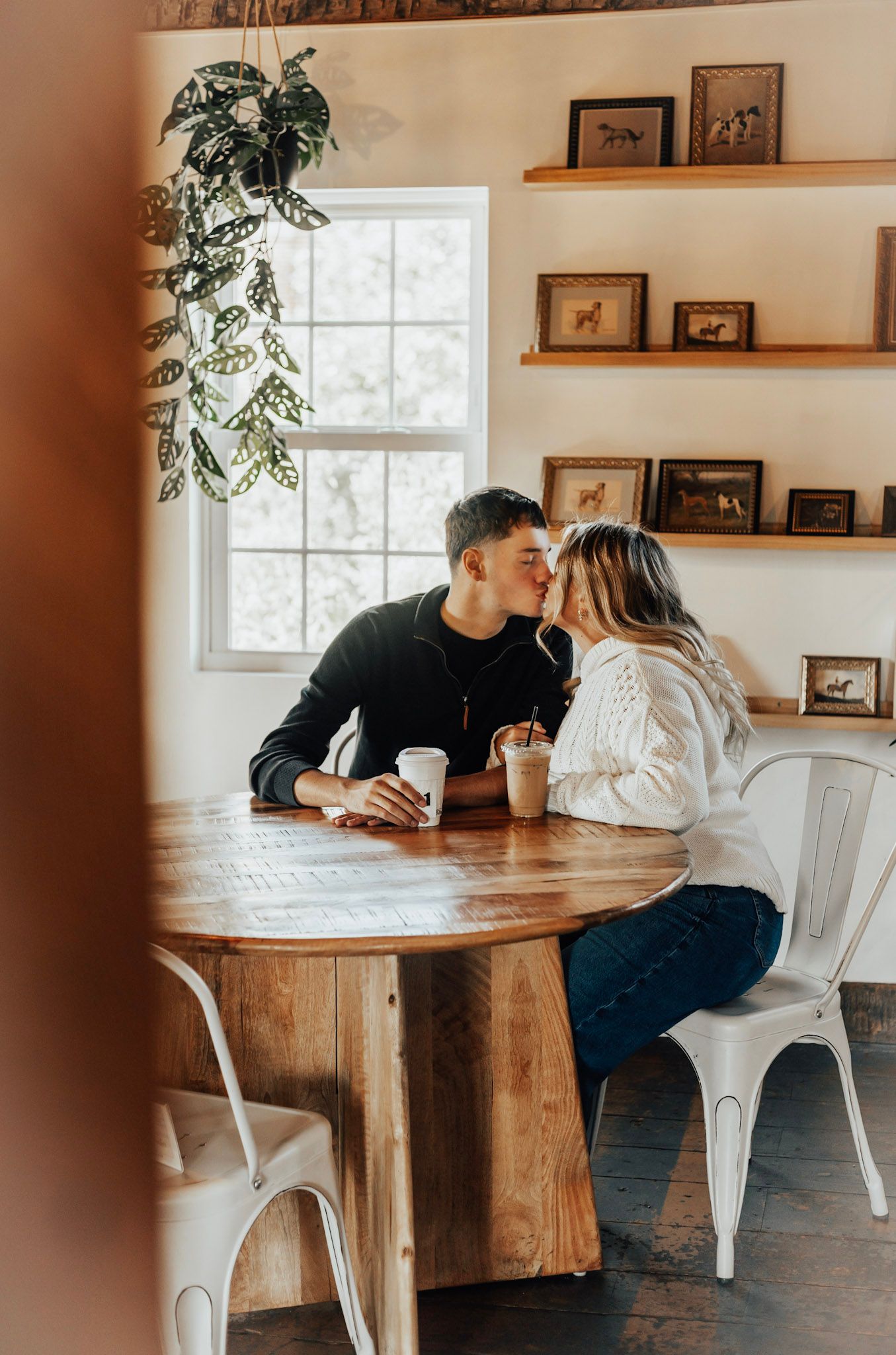 Couple sharing intimate moment at wooden dining table with white chairs and framed photos on wall.