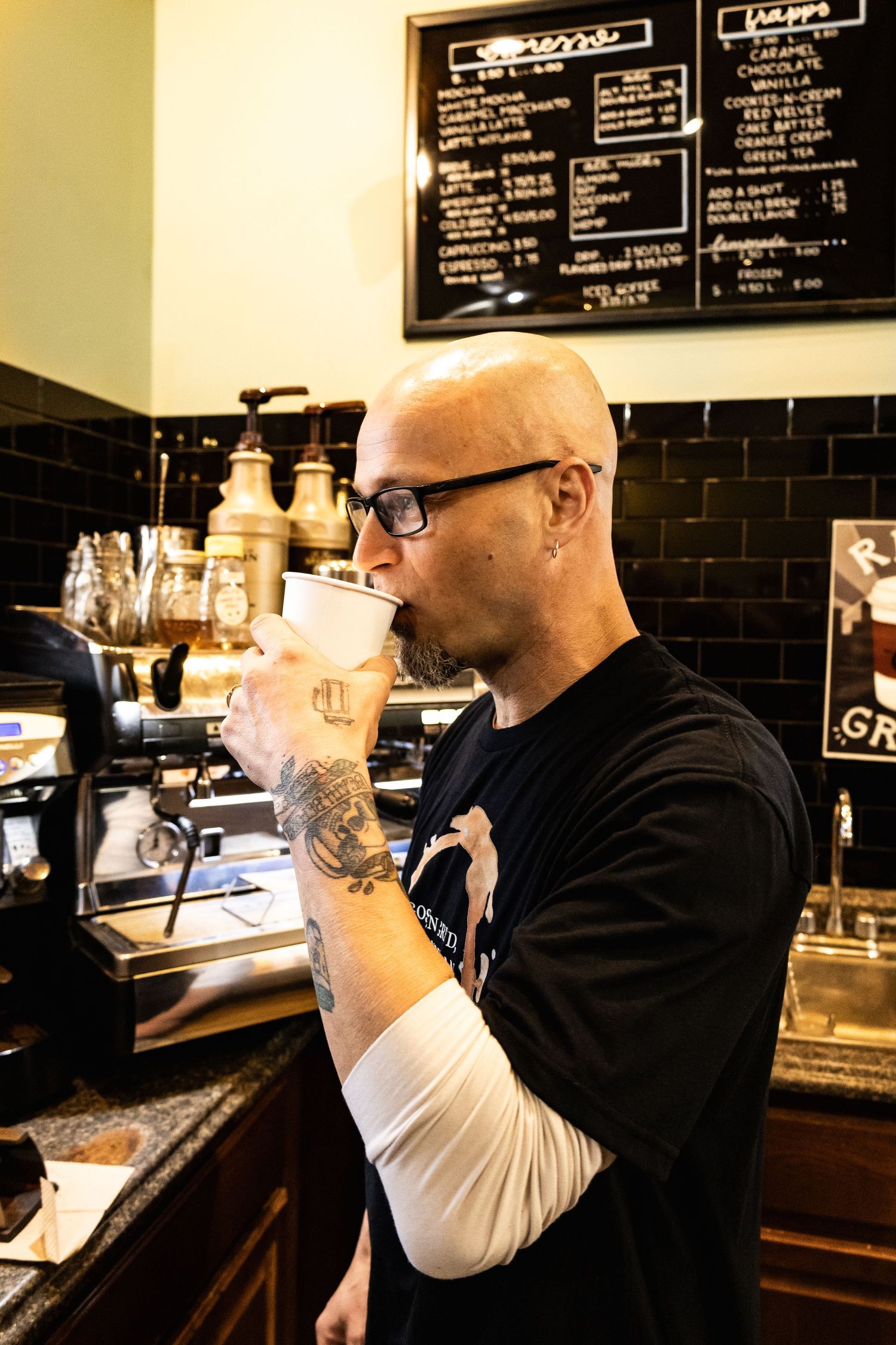Barista in black shirt tasting coffee at a cafe counter with menu boards visible.