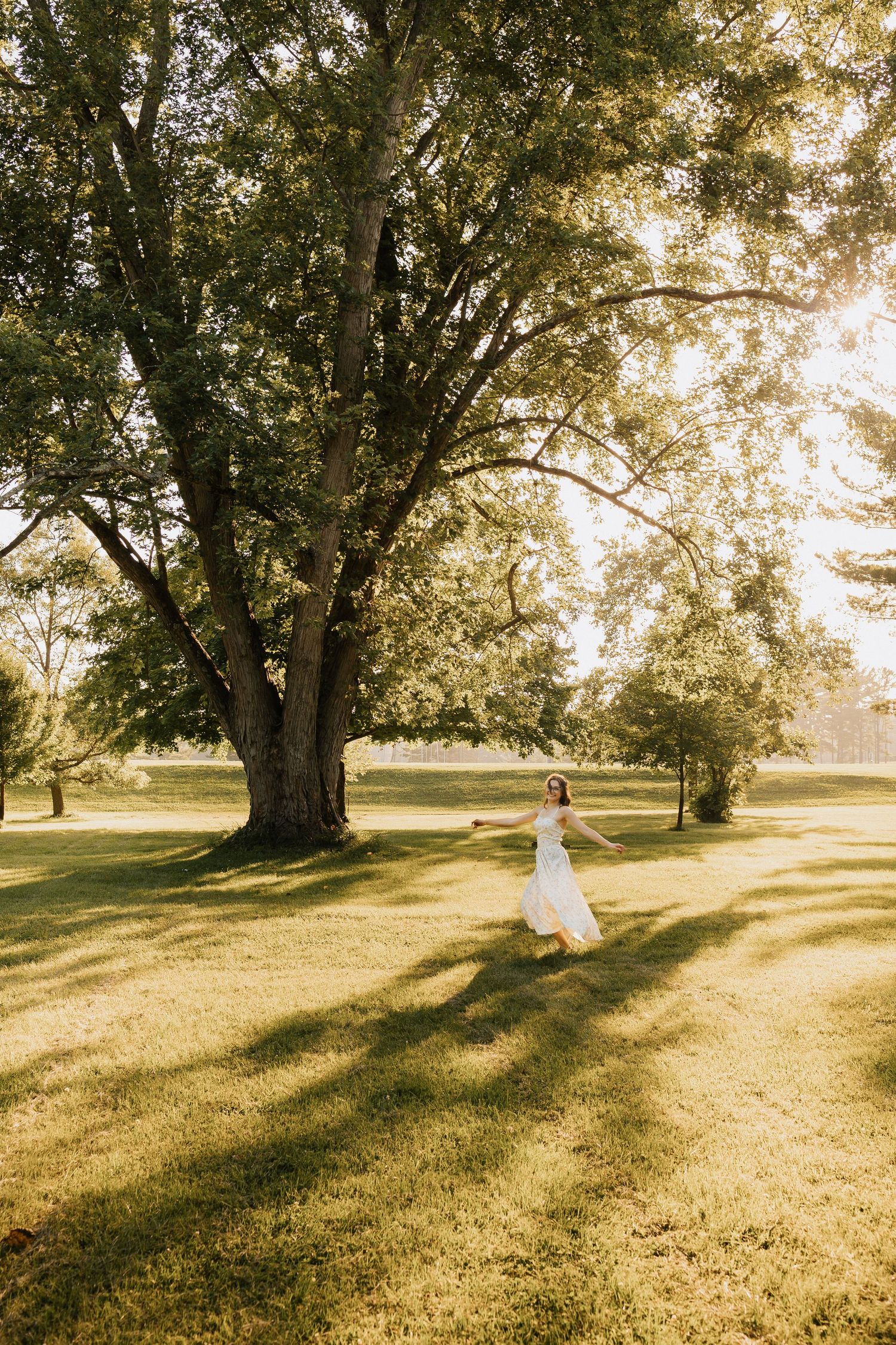 A bride in a white gown twirls gracefully under a large tree in a sunlit green park.