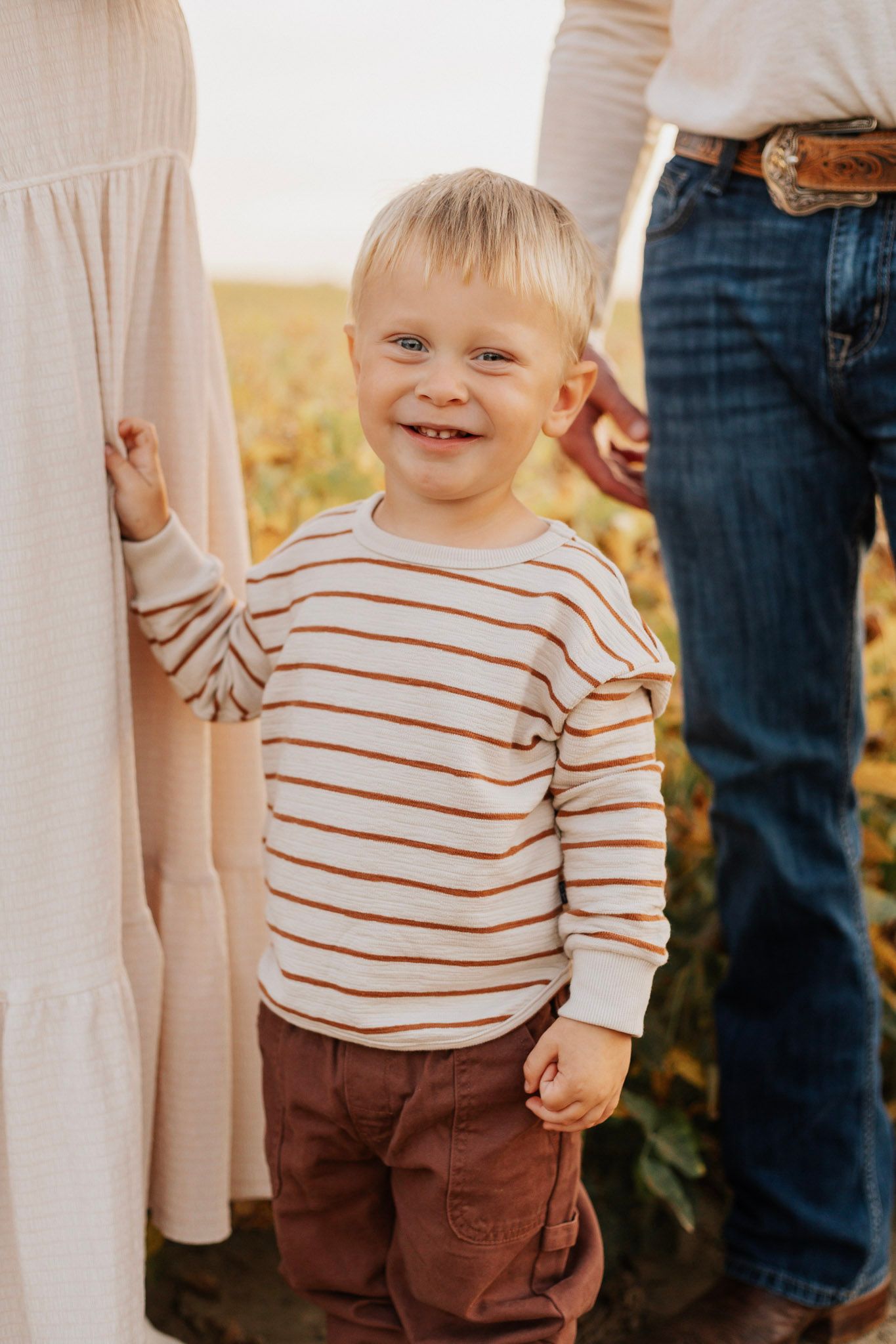 Child in striped shirt and brown pants smiling outdoors.