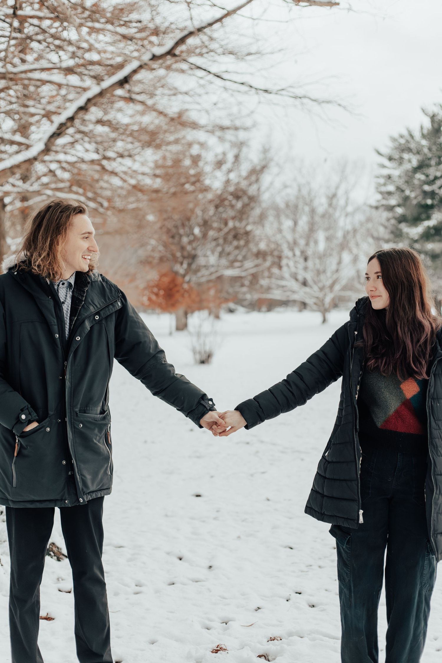 A couple holds hands while walking through a snow-covered path in the woods.