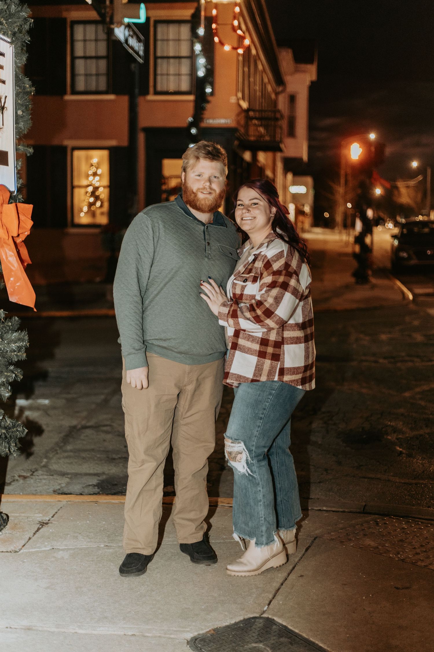 A couple poses together on a city sidewalk at night with string lights in the background.