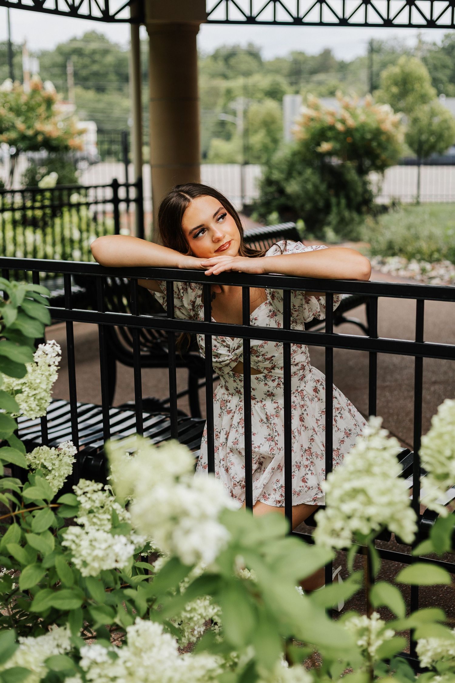 Woman in floral dress leans on railing surrounded by white hydrangea flowers.