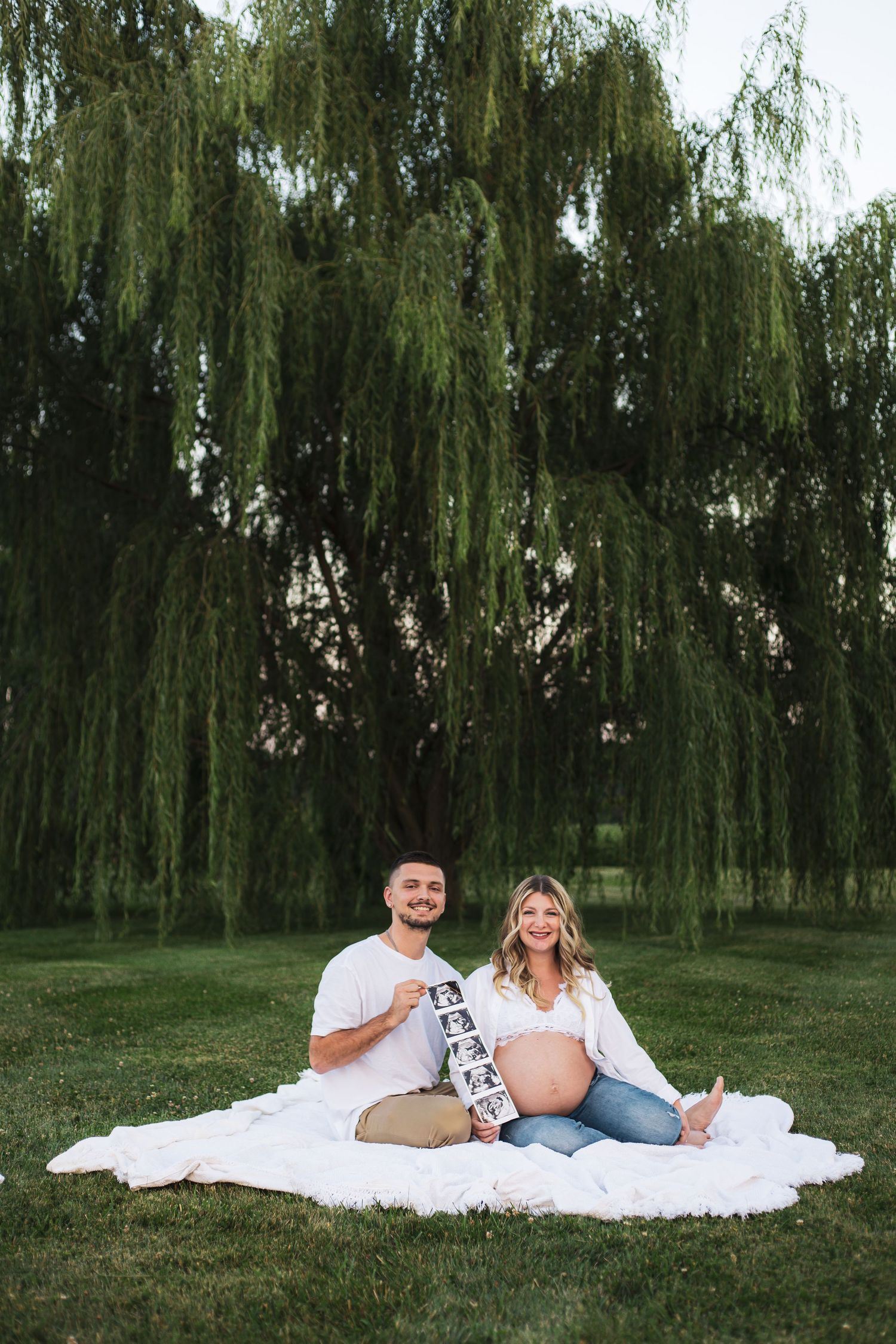 Expecting couple relaxing on white blanket under willow trees during outdoor photoshoot.