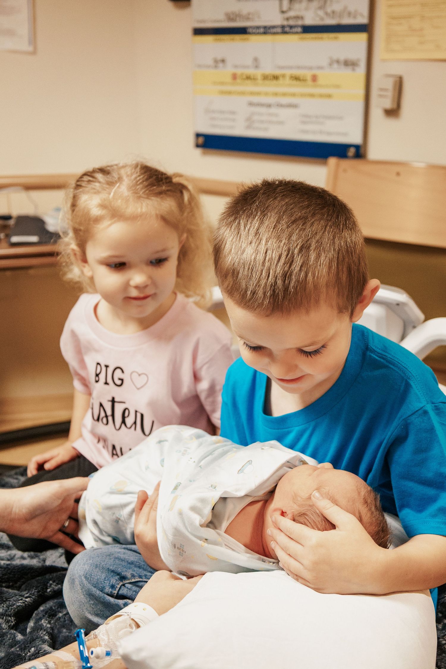 Children in blue and pink shirts meeting their new sibling in hospital.