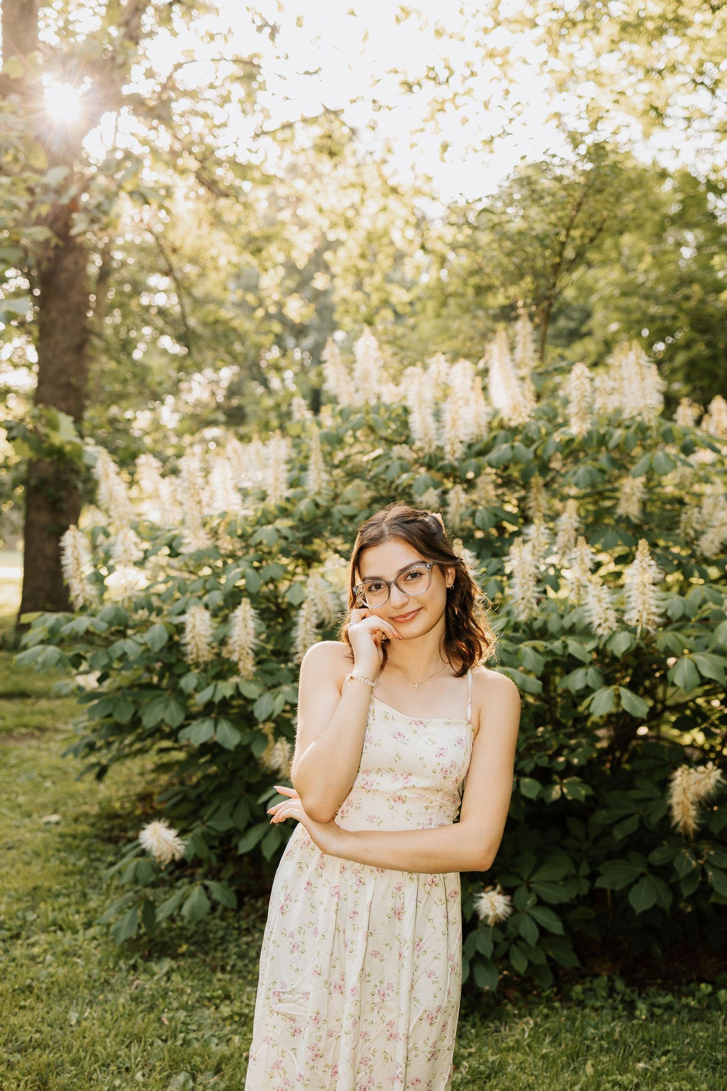 Young woman in floral dress and glasses poses before blooming white hydrangeas in a sunlit park.