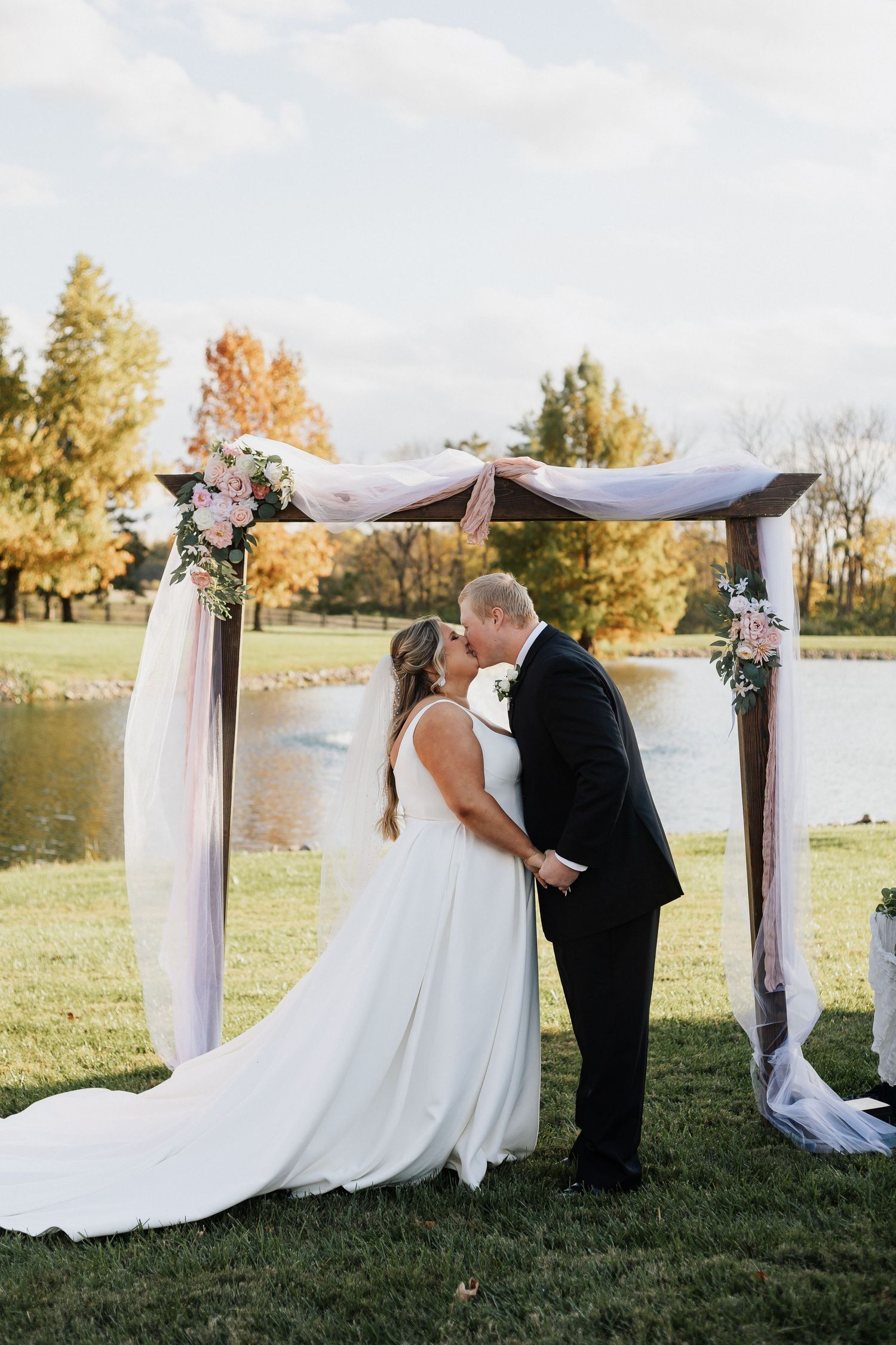 Couple shares intimate moment under draped wedding arch by lakeside during autumn ceremony.