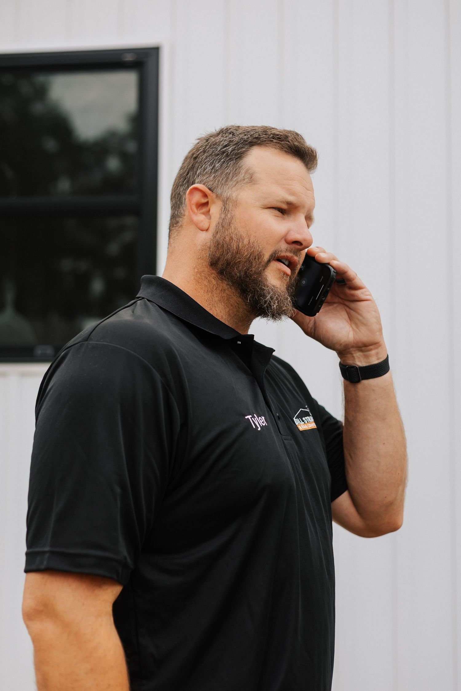 Person in black polo shirt talking on mobile phone outside white building.