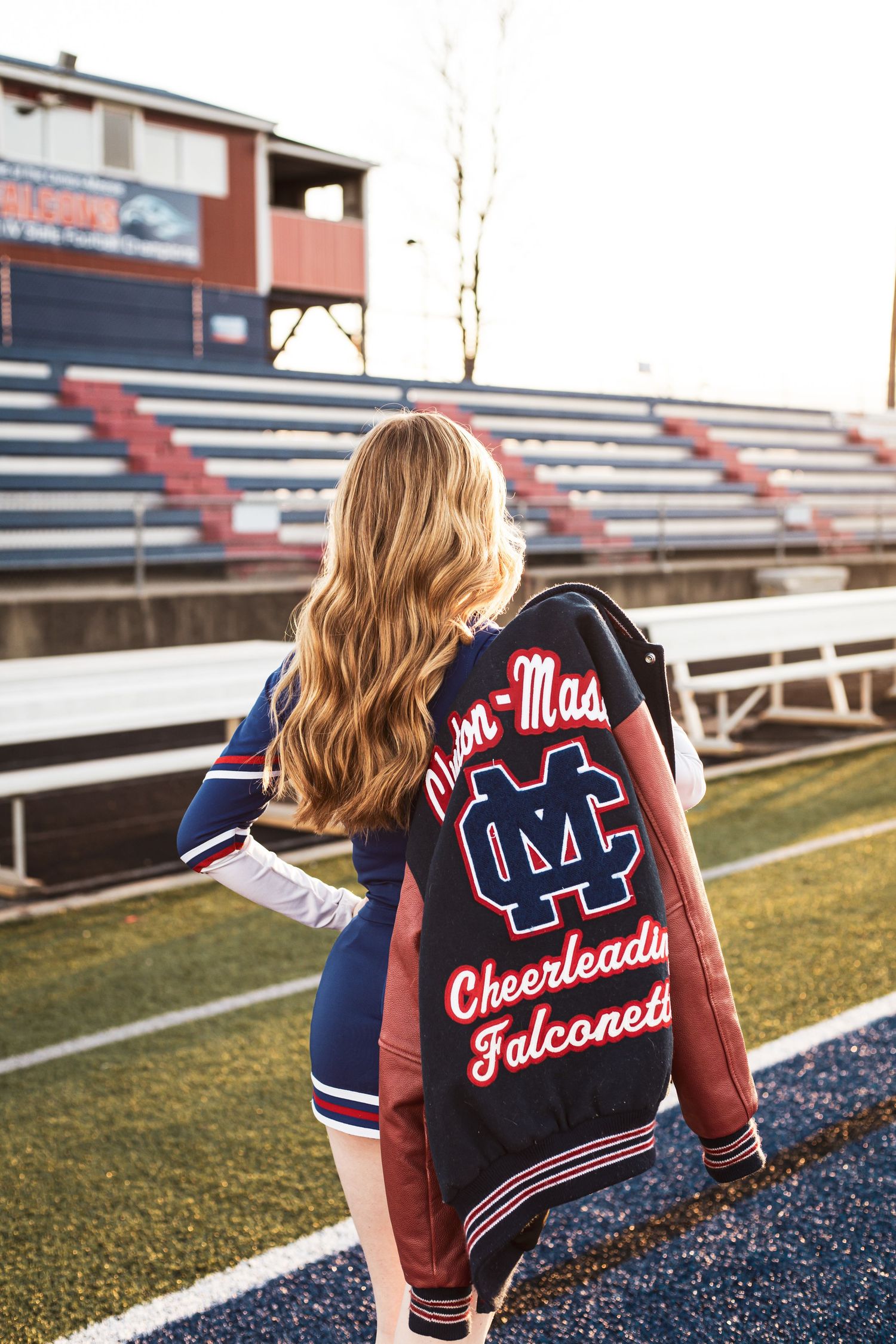Person wearing Ole Miss Cheerleader Falcons varsity jacket on football field with bleachers.