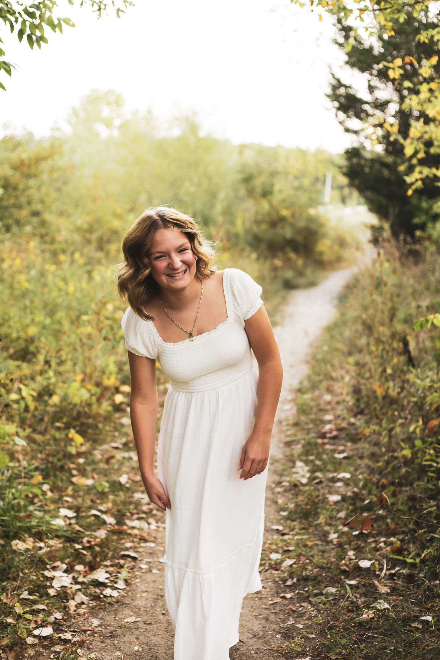 Person in flowing white dress walking along nature path with golden sunlight.