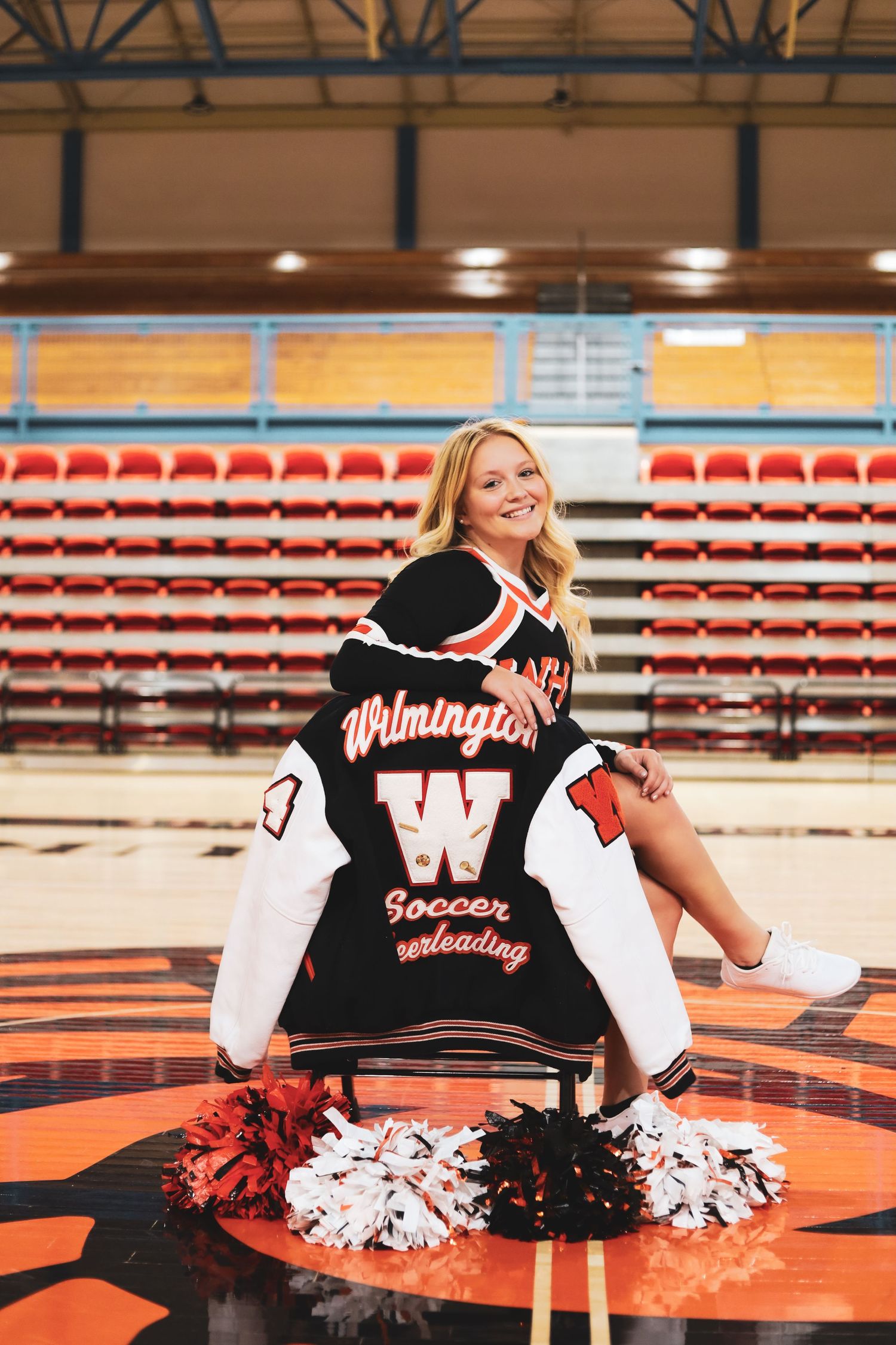 High school cheerleader with varsity jacket and pom poms posing in a gymnasium with stadium seating.