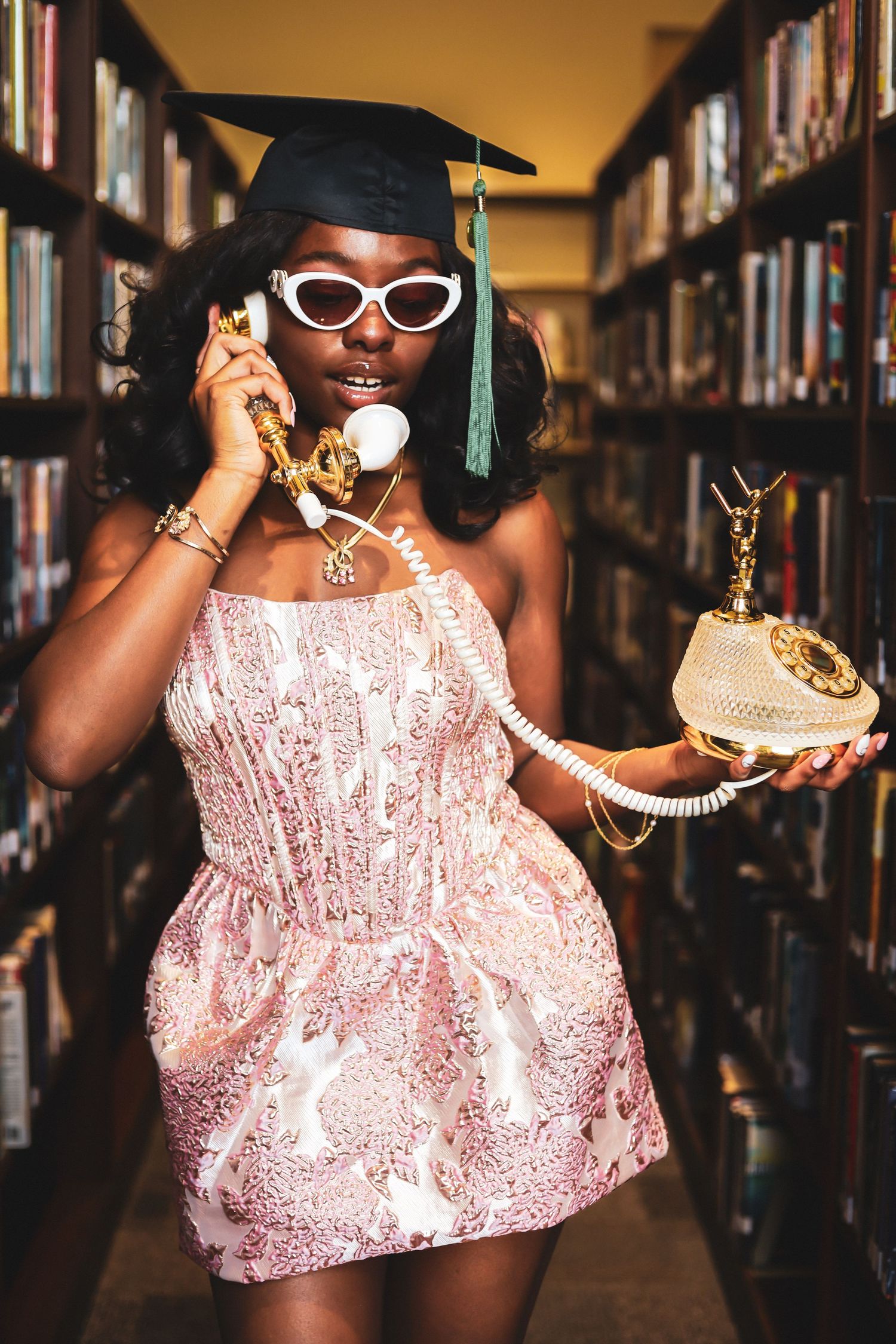 Graduate in sparkly pink dress poses with vintage telephone between library bookshelves.