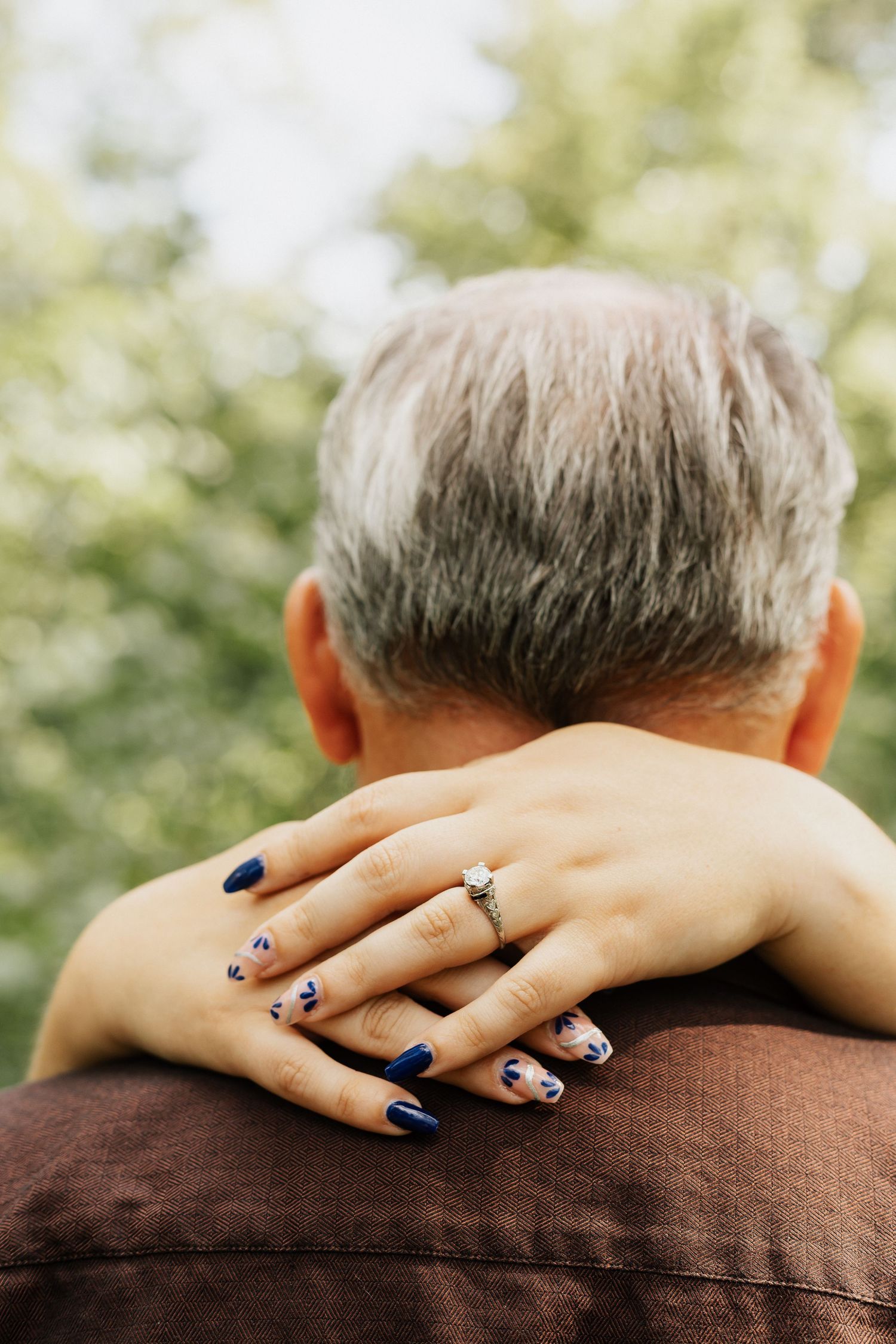 Close-up of hands with blue nail polish and engagement ring resting on brown fabric.