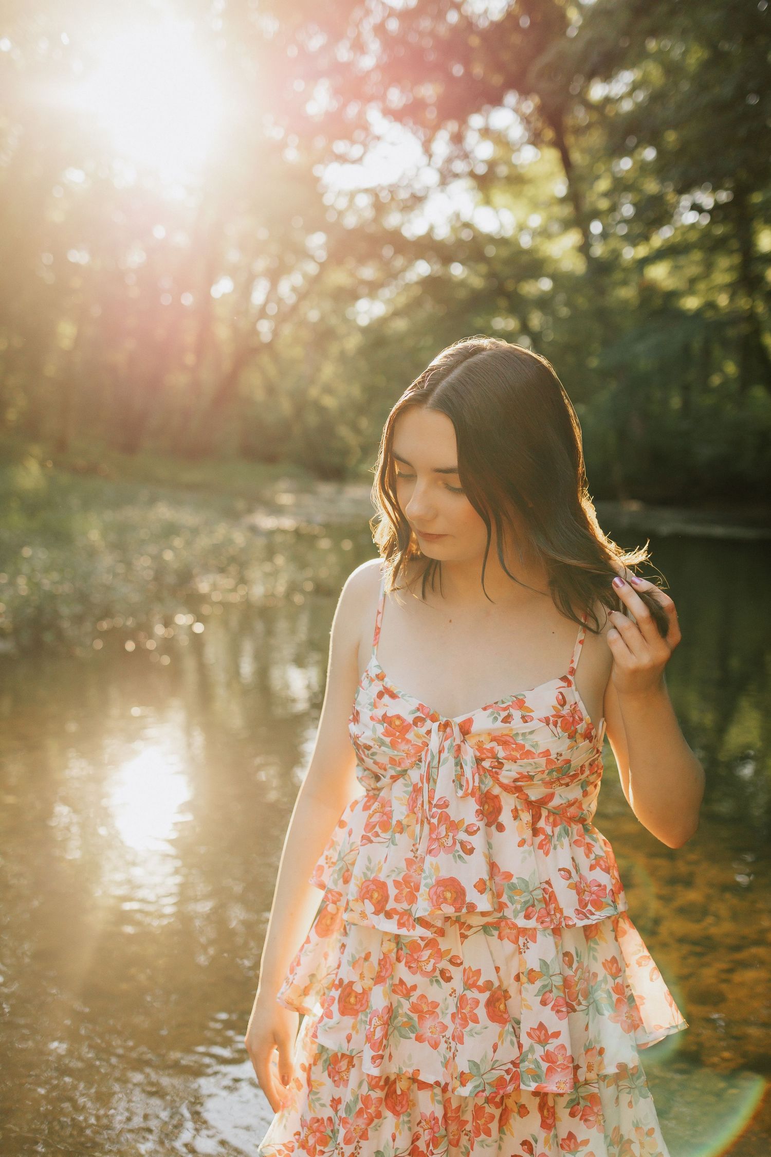 Young woman in floral sundress standing by a sunlit forest creek, golden hour light glowing behind her.