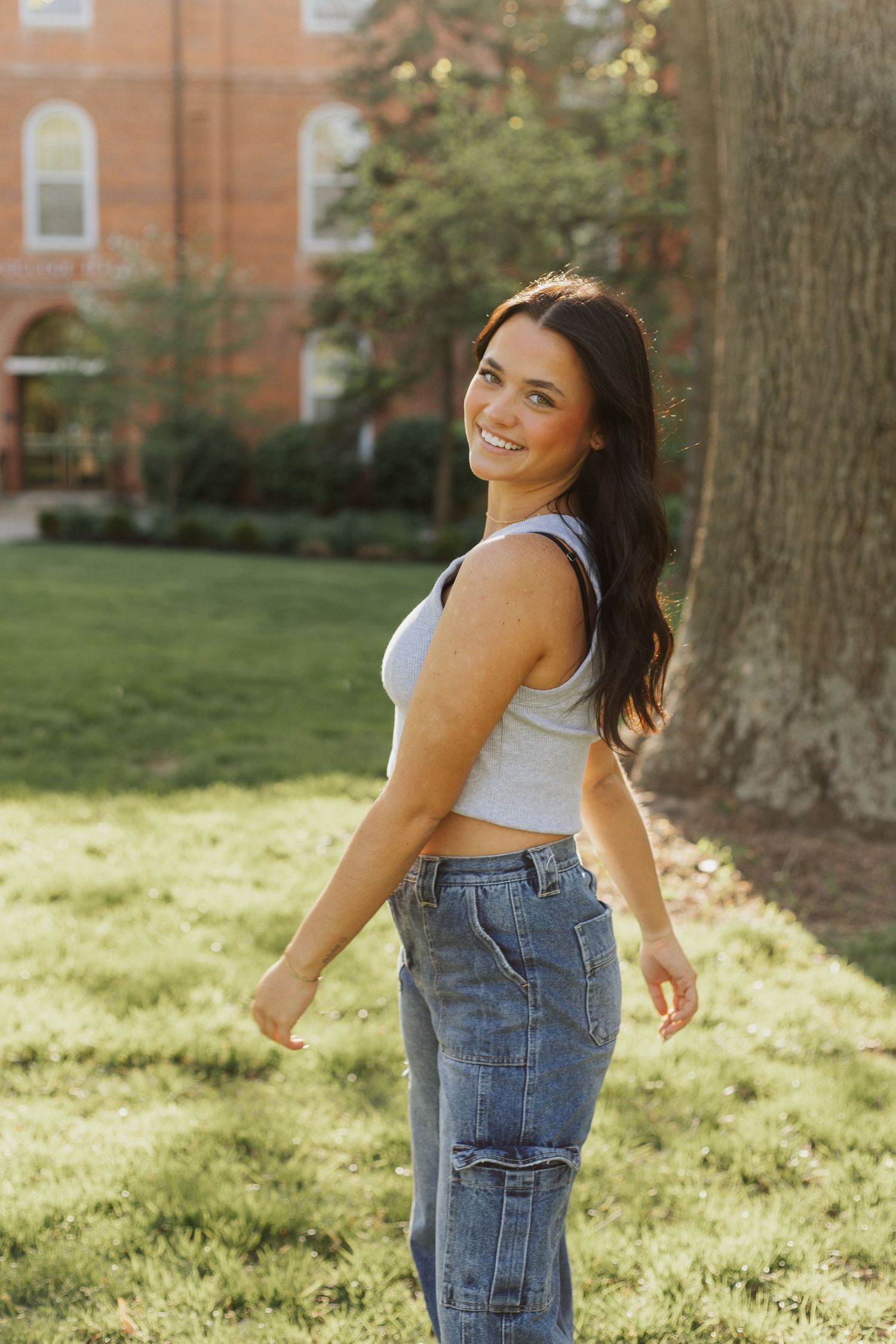 Person in white tank top and cargo jeans poses on sunny campus lawn.