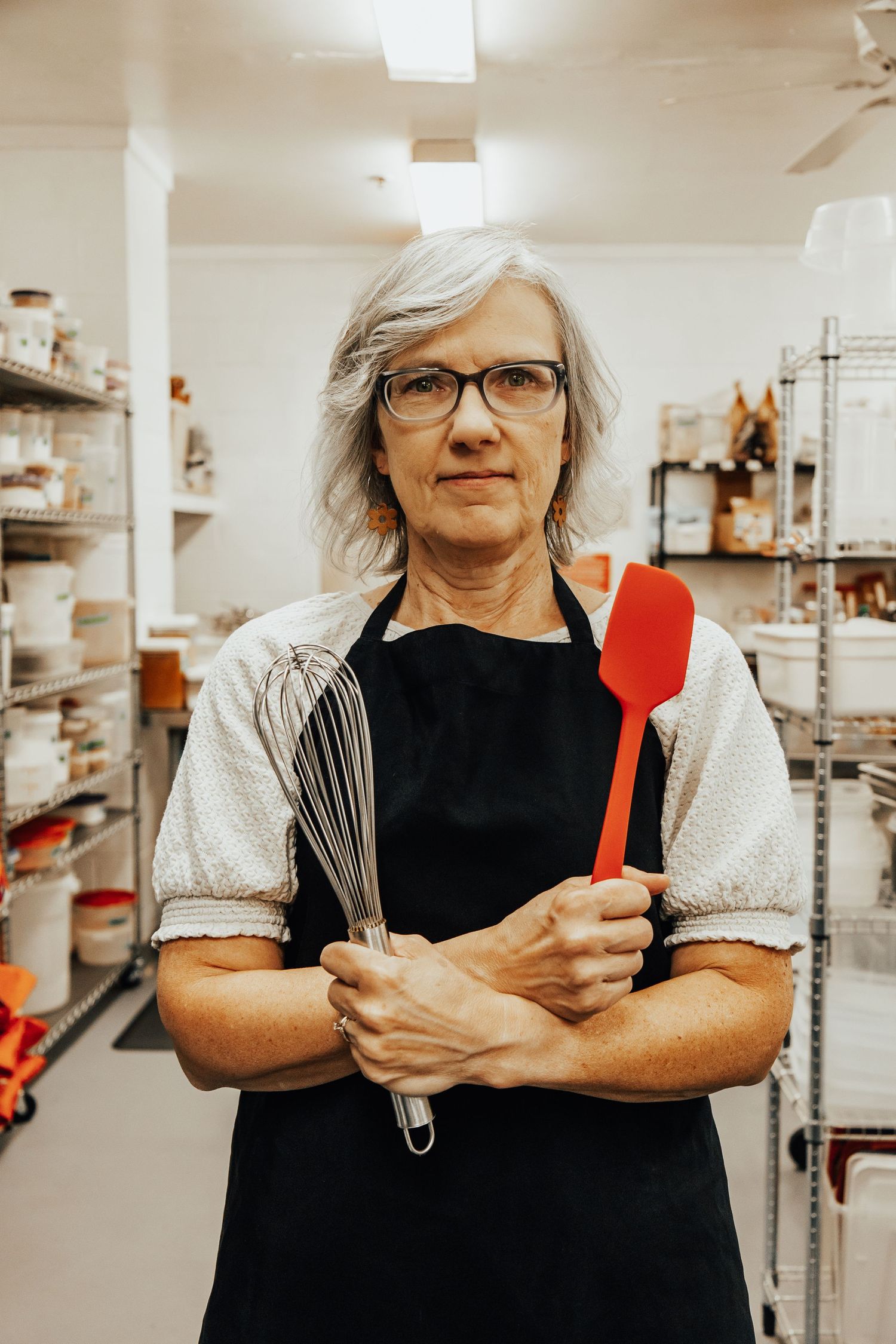 Baker wearing black apron and glasses holding red kitchen utensils in a commercial kitchen.