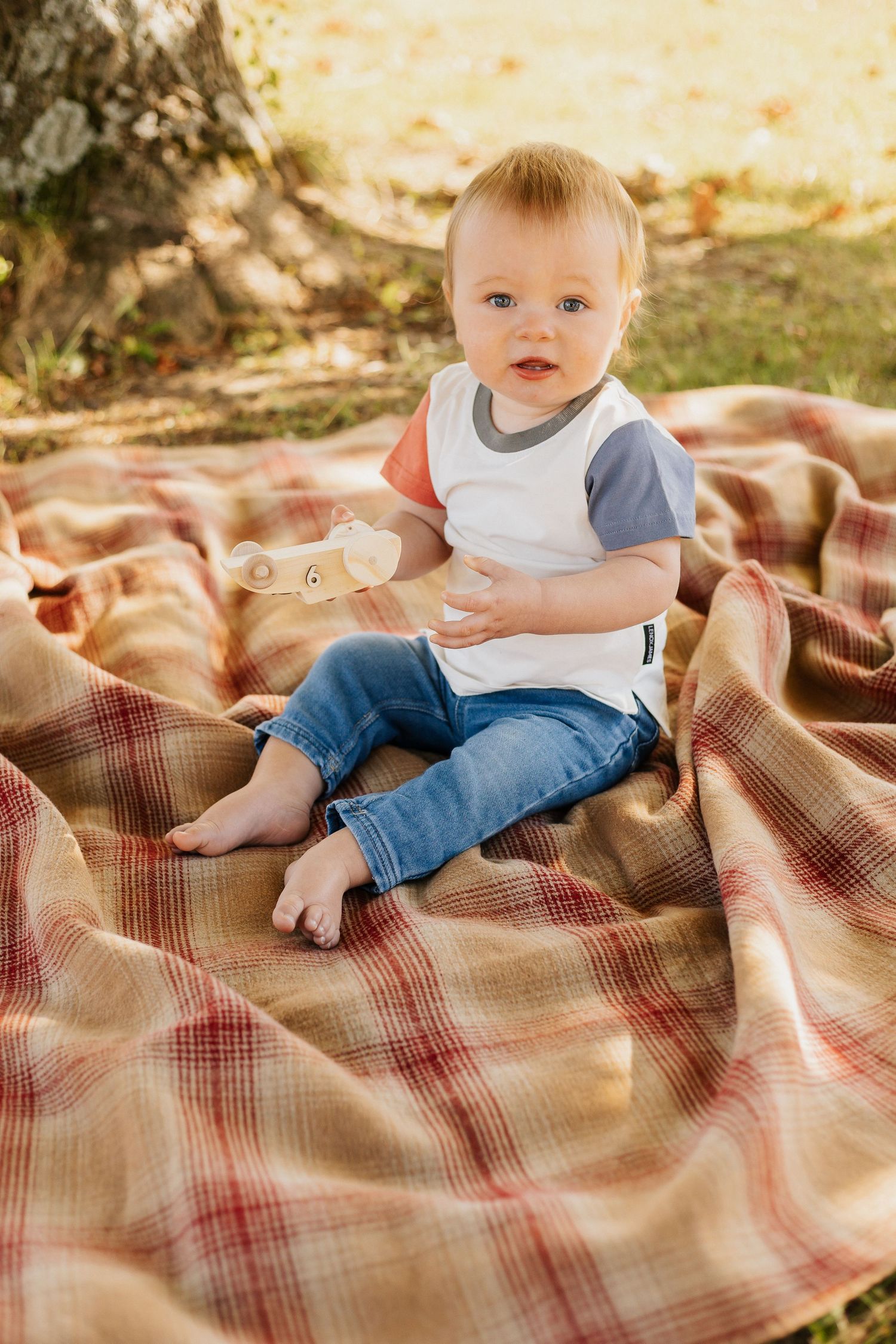Baby in white and gray t-shirt sitting on plaid blanket outdoors.