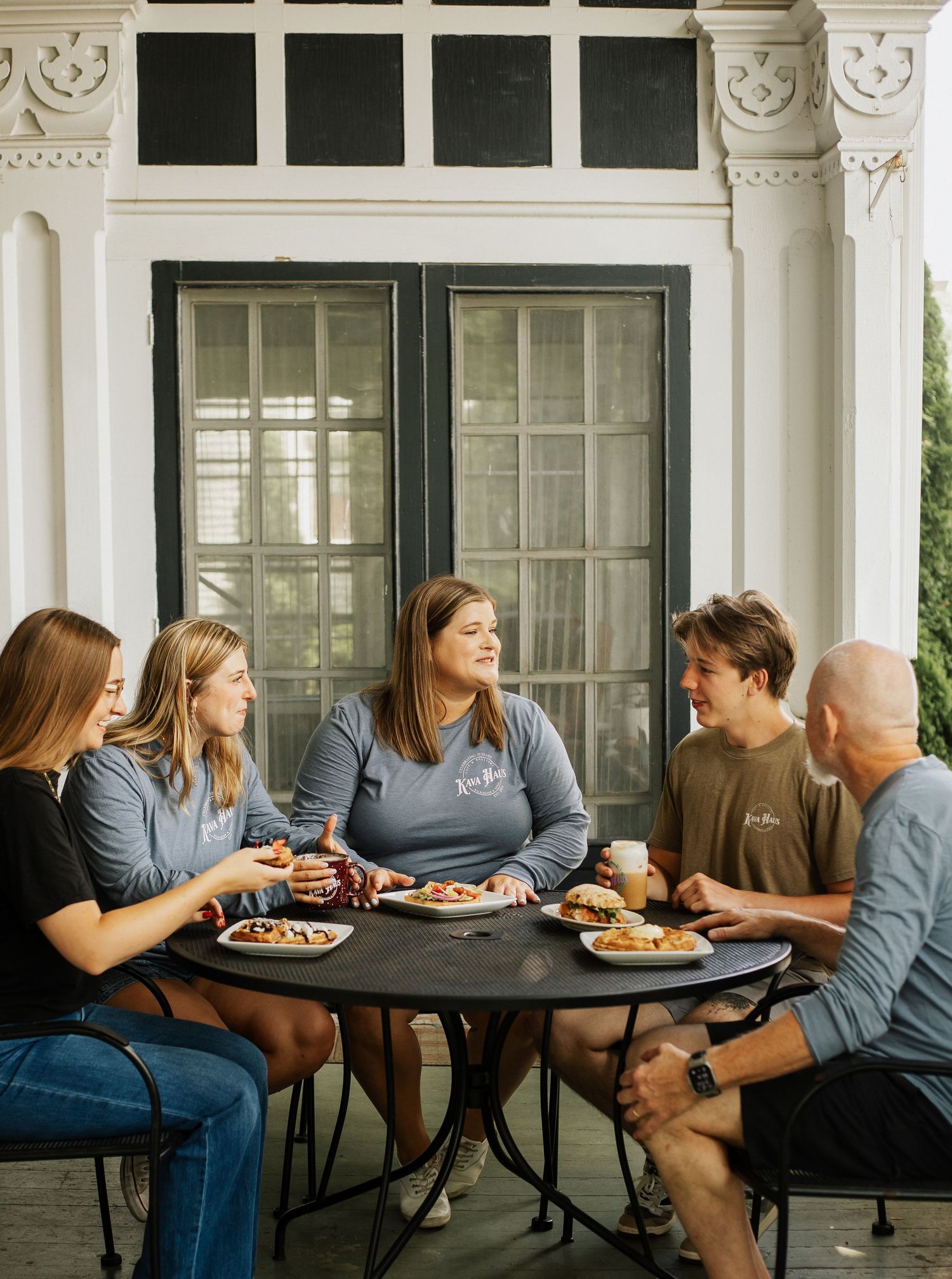 Group enjoying a meal together at a round table on a sunny porch.