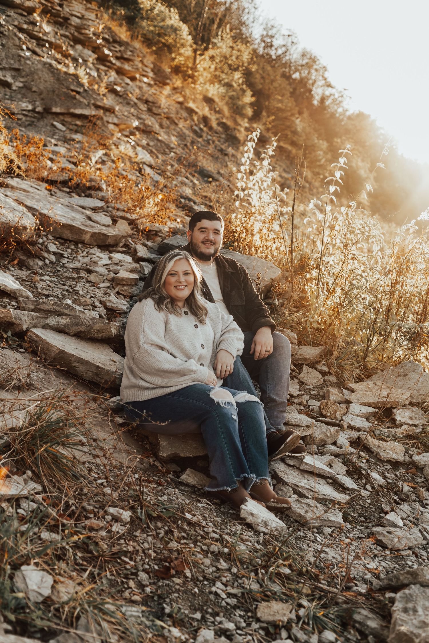 Couple sitting together on rocky cliff during golden hour sunset in autumn landscape.