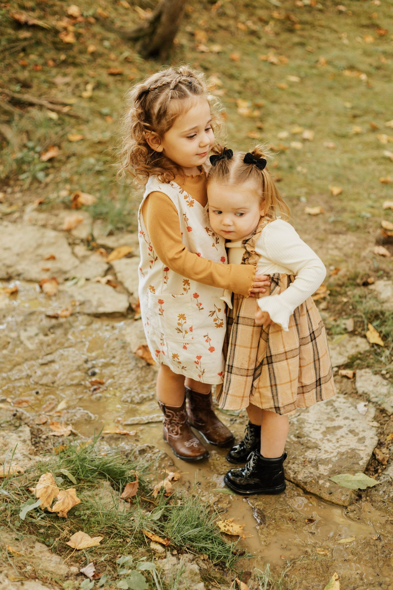 Two young children in vintage-style clothing playing together on rocky ground with autumn leaves.